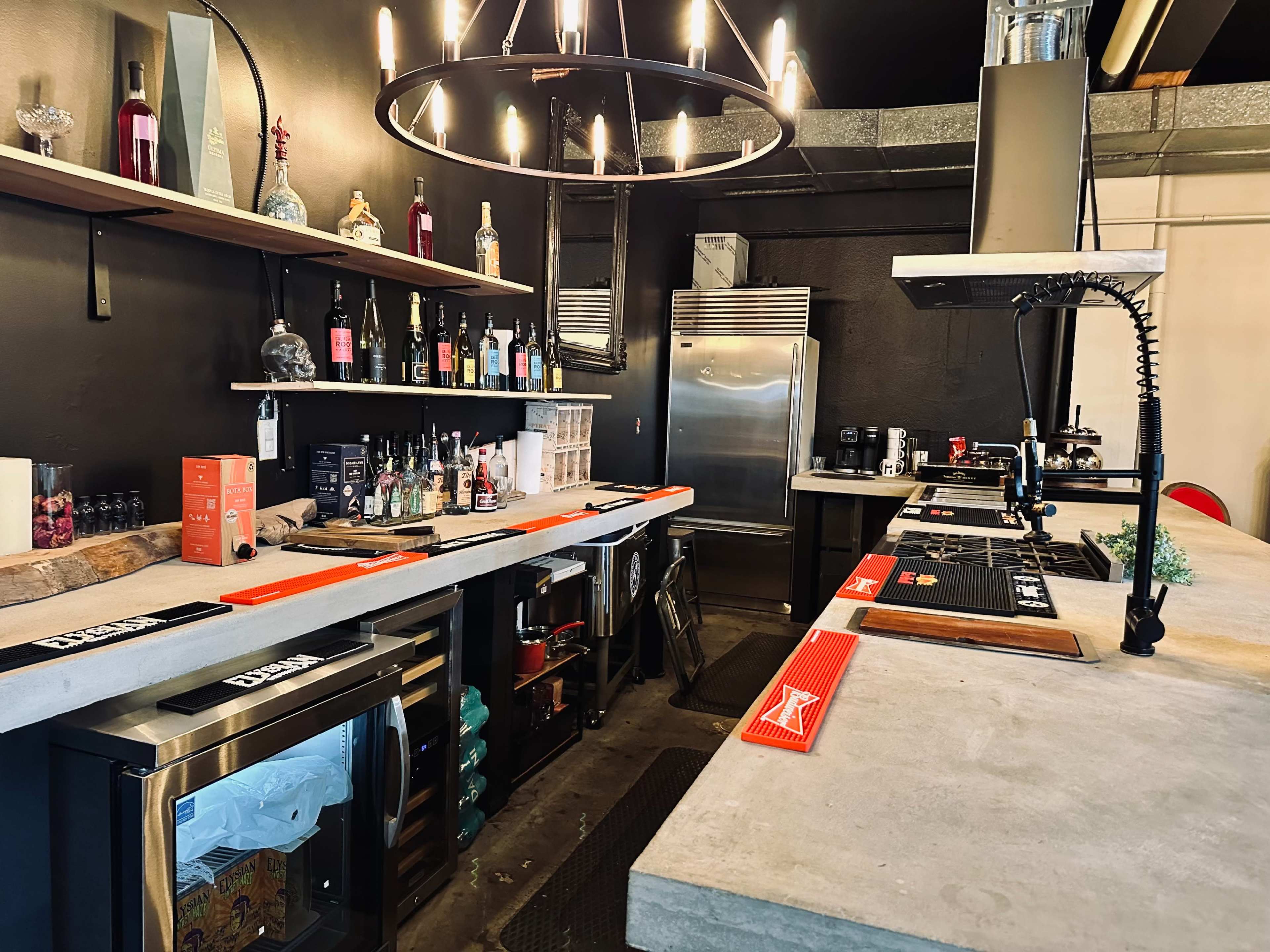 The image shows a modern bar setup featuring shelves stocked with various bottles, a stainless steel refrigerator, and a concrete countertop with drink dispensers and glassware.