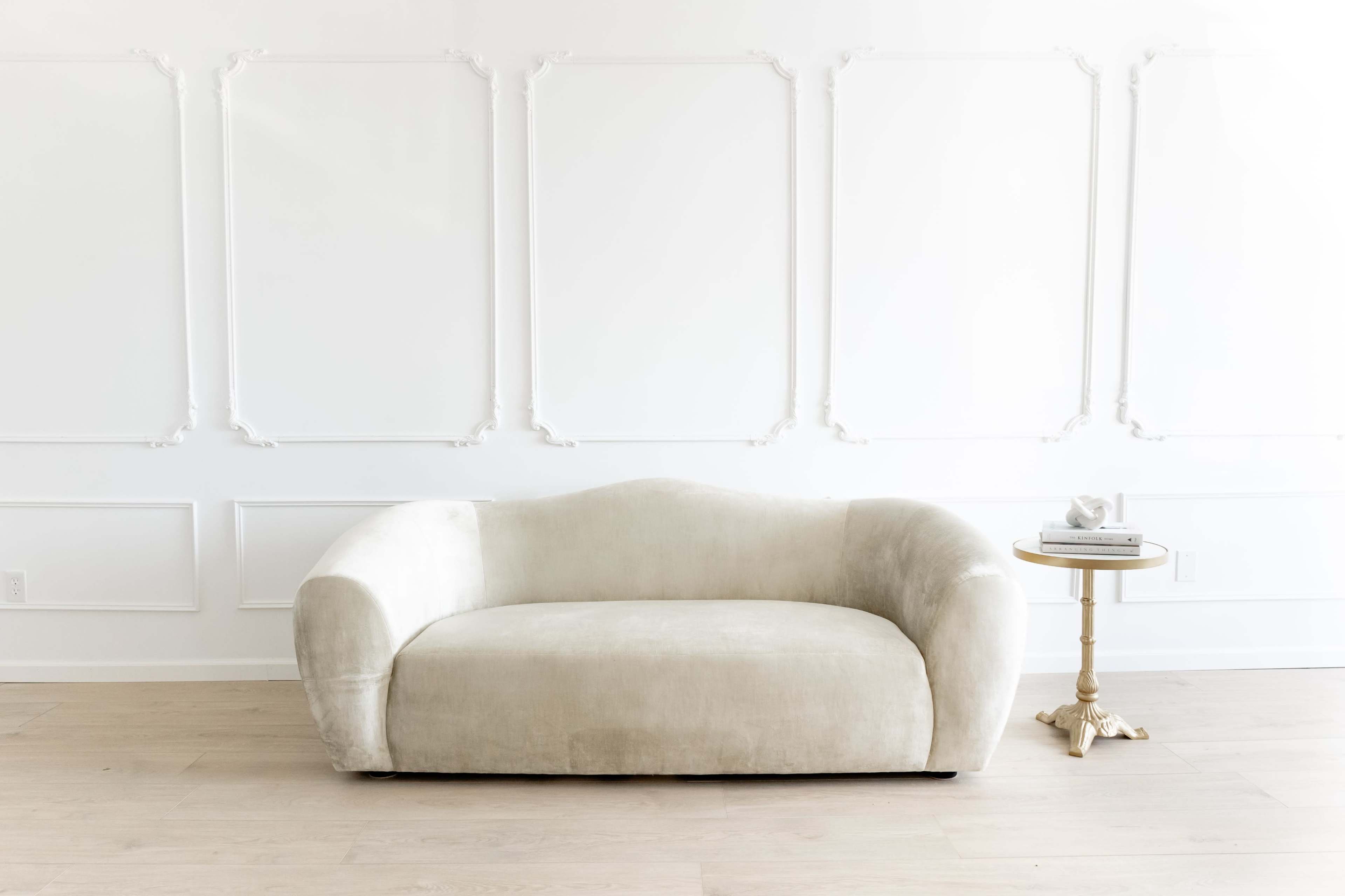 A beige curved sofa is positioned in front of a white wall with decorative panels, next to a small round table.