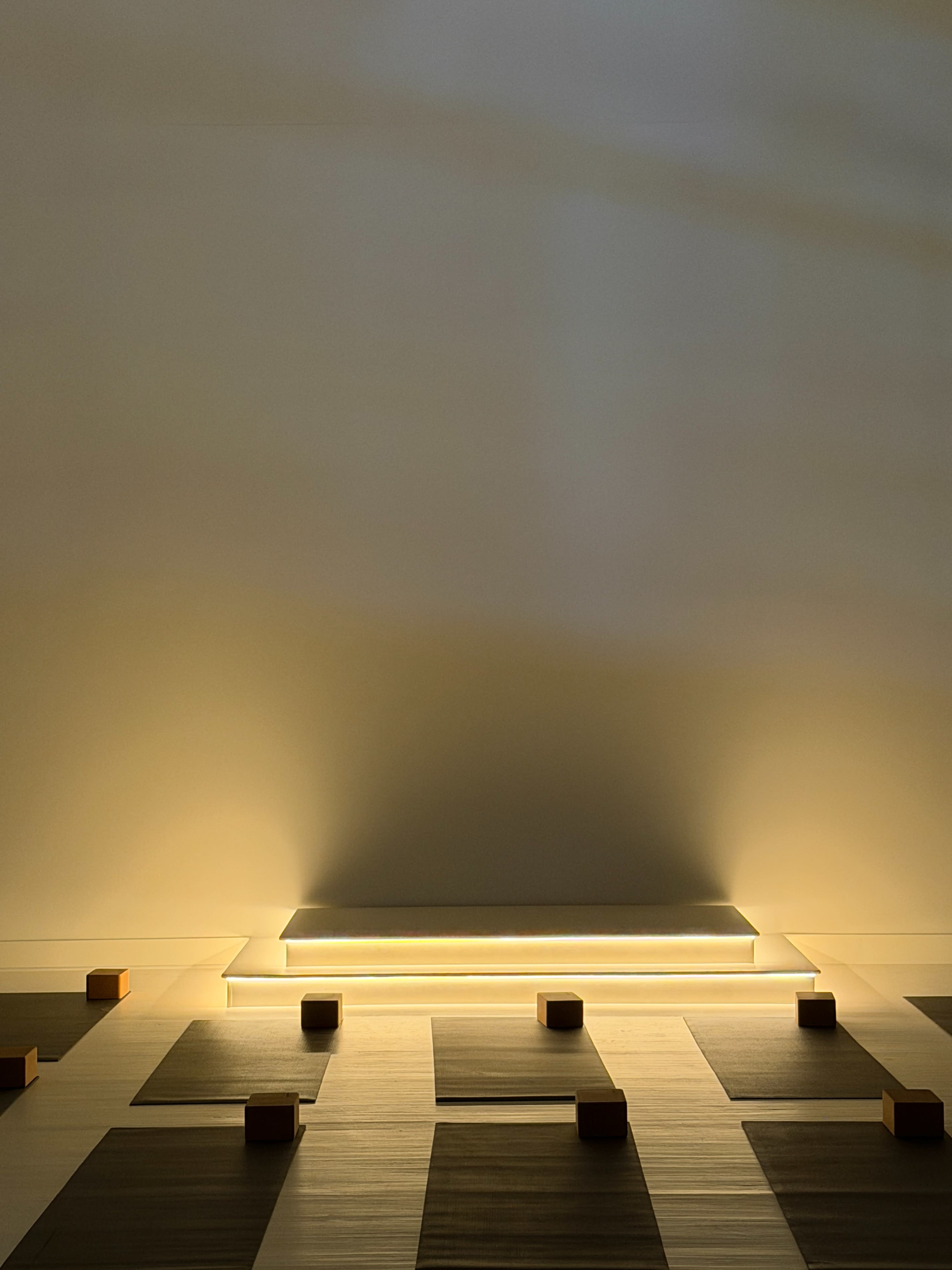 The image shows a minimalist room with a softly illuminated platform and scattered wooden cubes on a wooden floor.