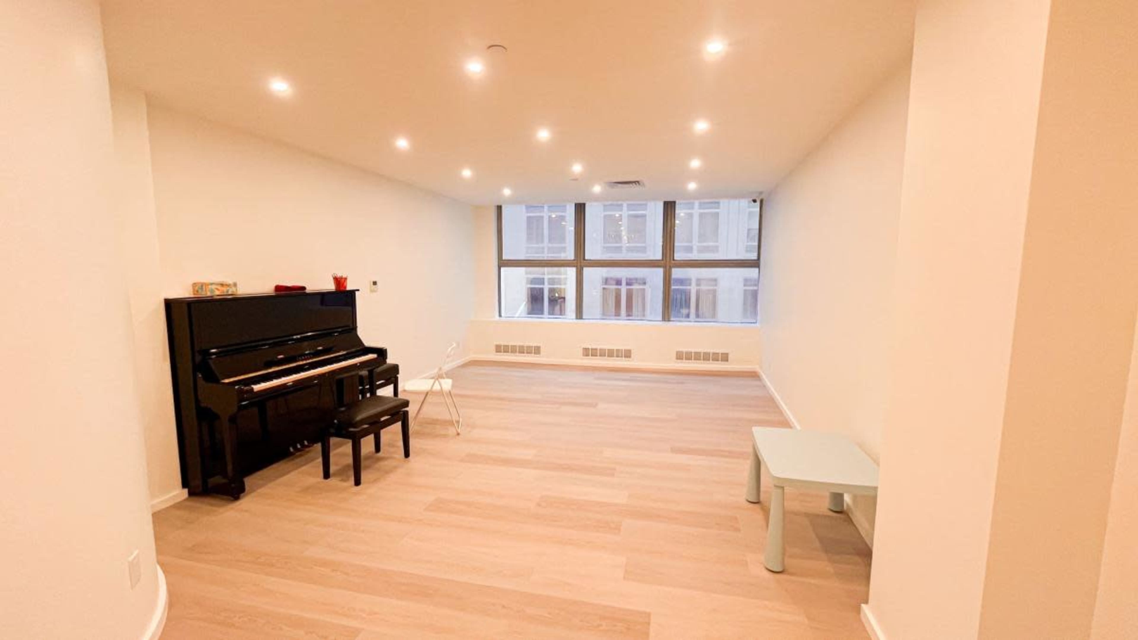 The image shows a spacious, empty room with a black piano, a chair, and a small light blue table, illuminated by recessed ceiling lights.