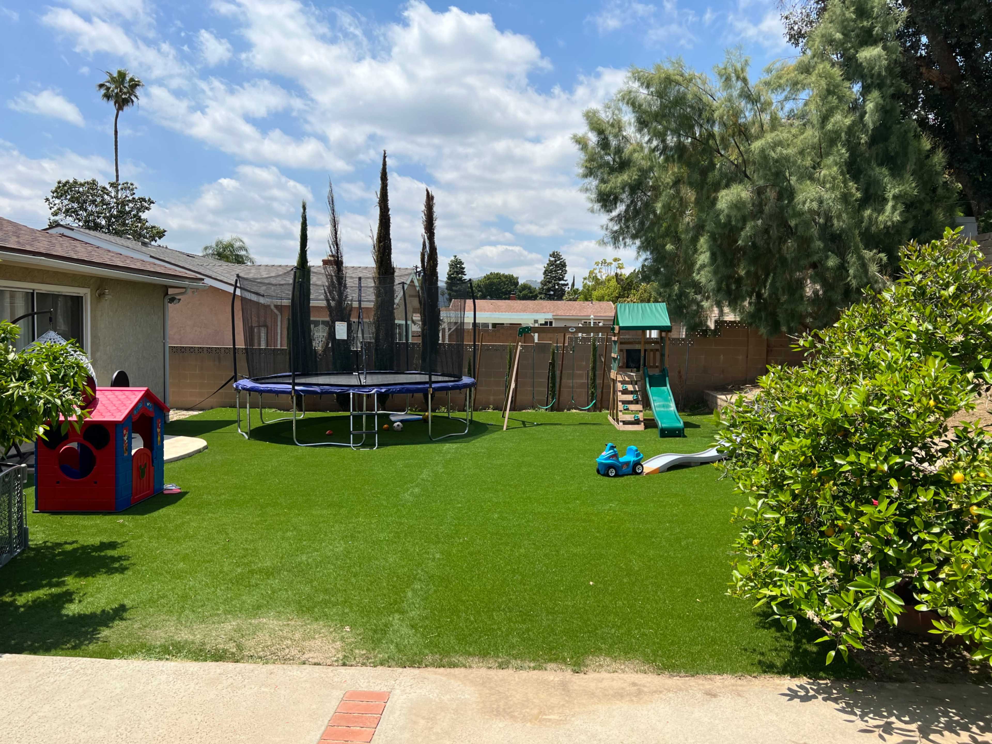 A backyard features a trampoline, a playground set, a red playhouse, and artificial grass.