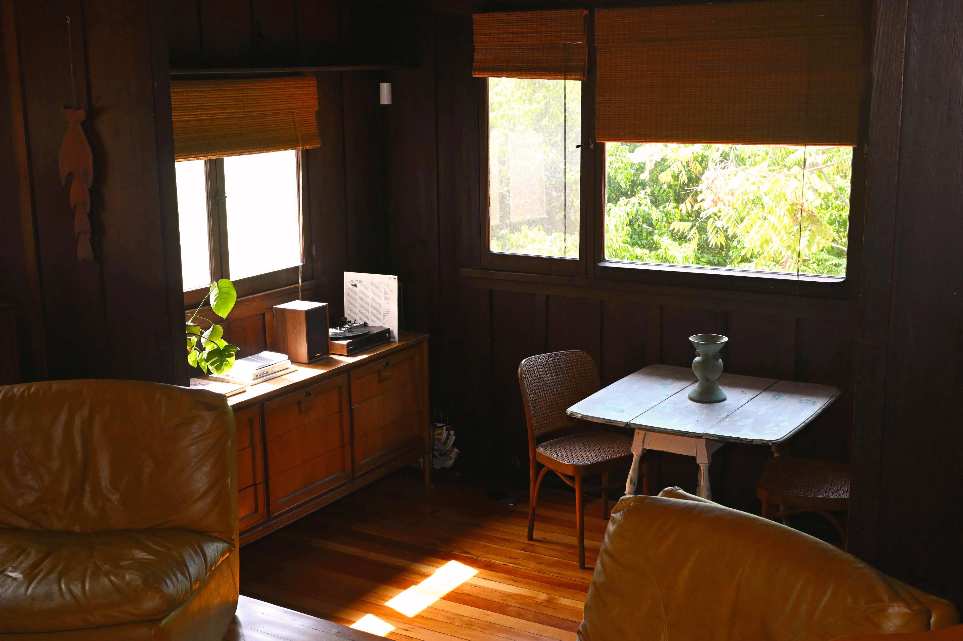 The image shows a cozy interior space featuring a wooden sideboard, a small table with chairs, and two large leather armchairs under natural light from the windows.