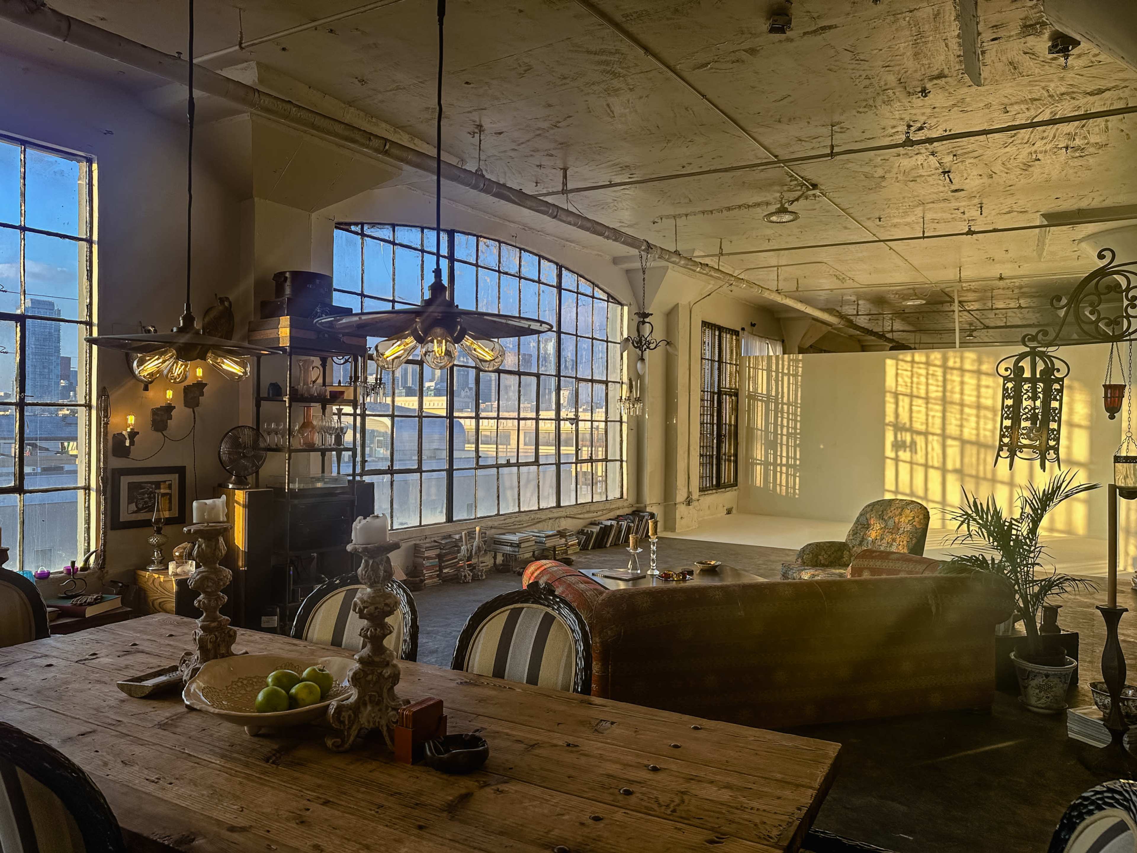 The image shows a spacious, well-lit loft with large windows, featuring a wooden dining table, a vintage-style couch, and various decorative elements against a backdrop of exposed brick and concrete walls.