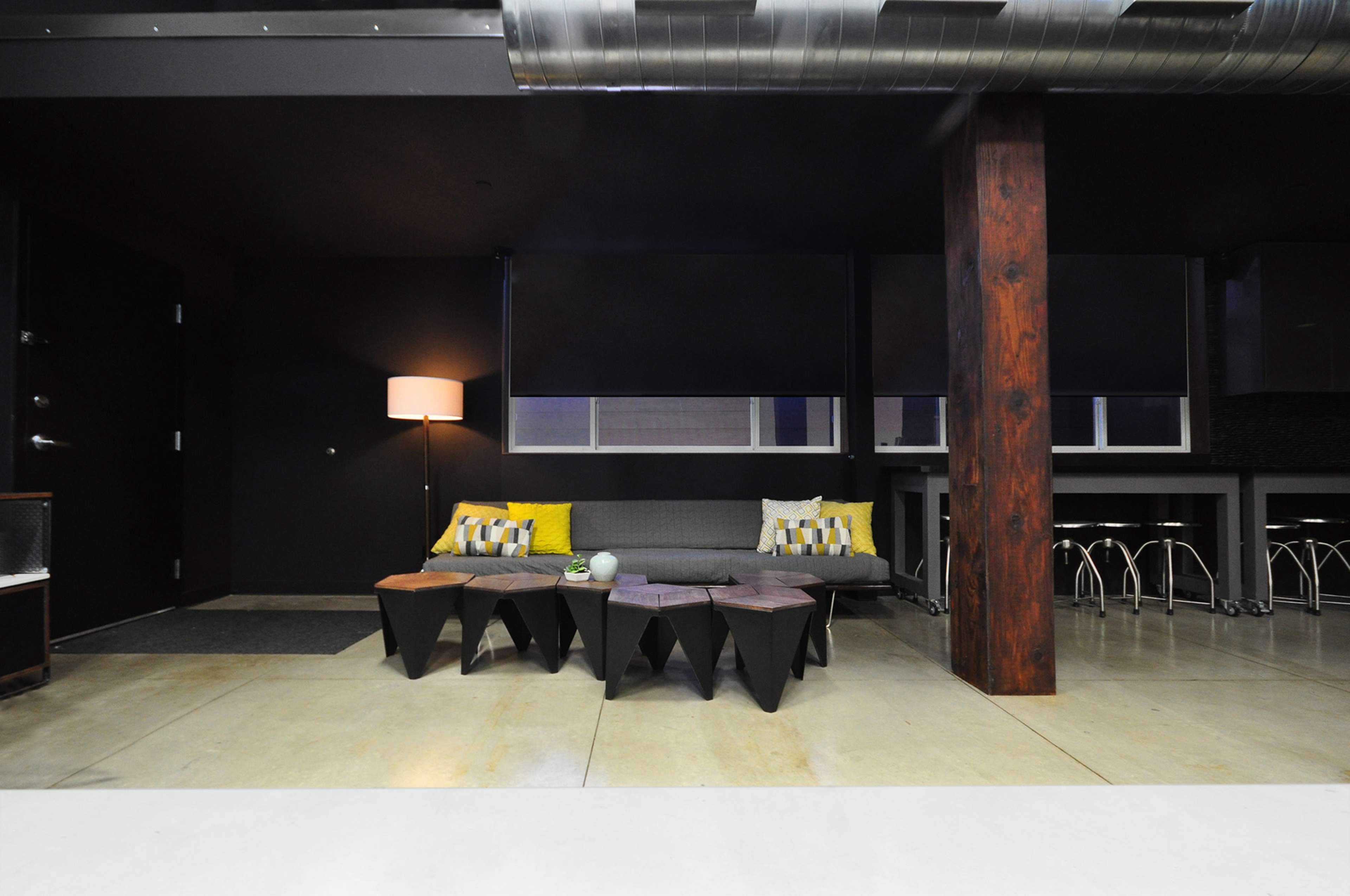 The image shows a modern, minimalist living area with a gray sofa, geometric black coffee tables, a floor lamp, and large windows.