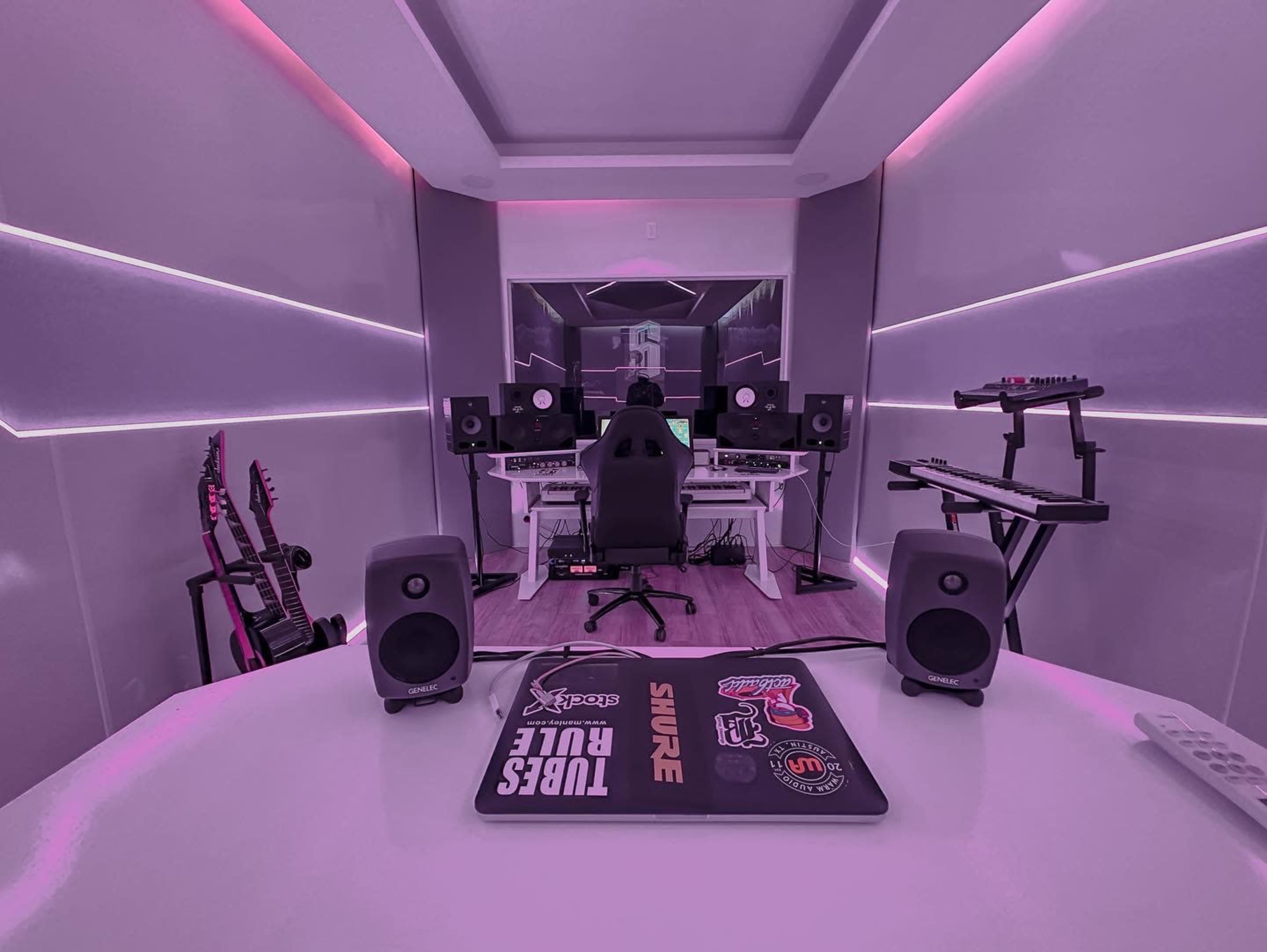 A music production studio features a white desk with speakers, a computer, and musical instruments, illuminated by purple lighting.