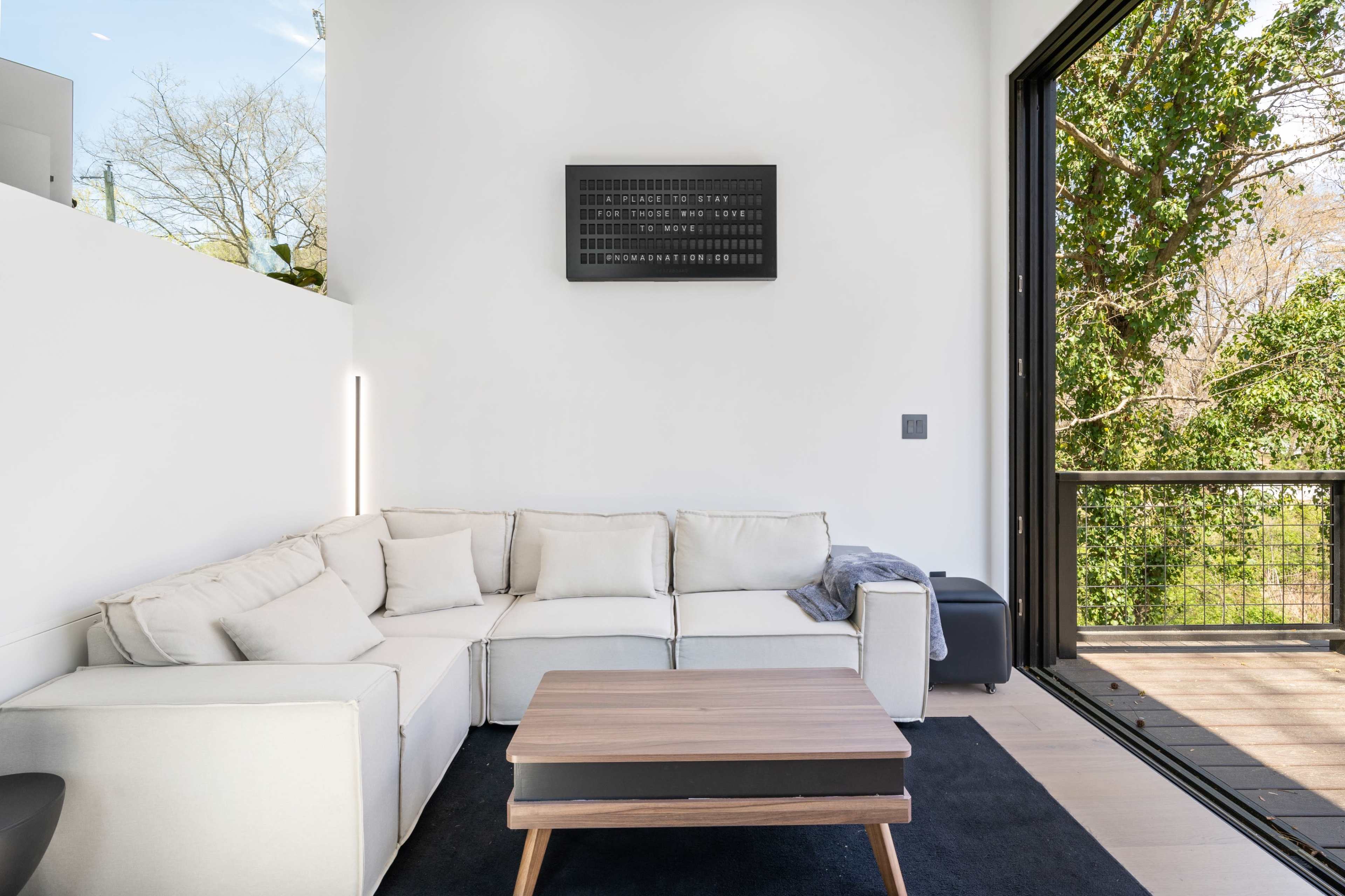 A modern living room features a white sectional sofa, a wooden coffee table, and large windows overlooking greenery.