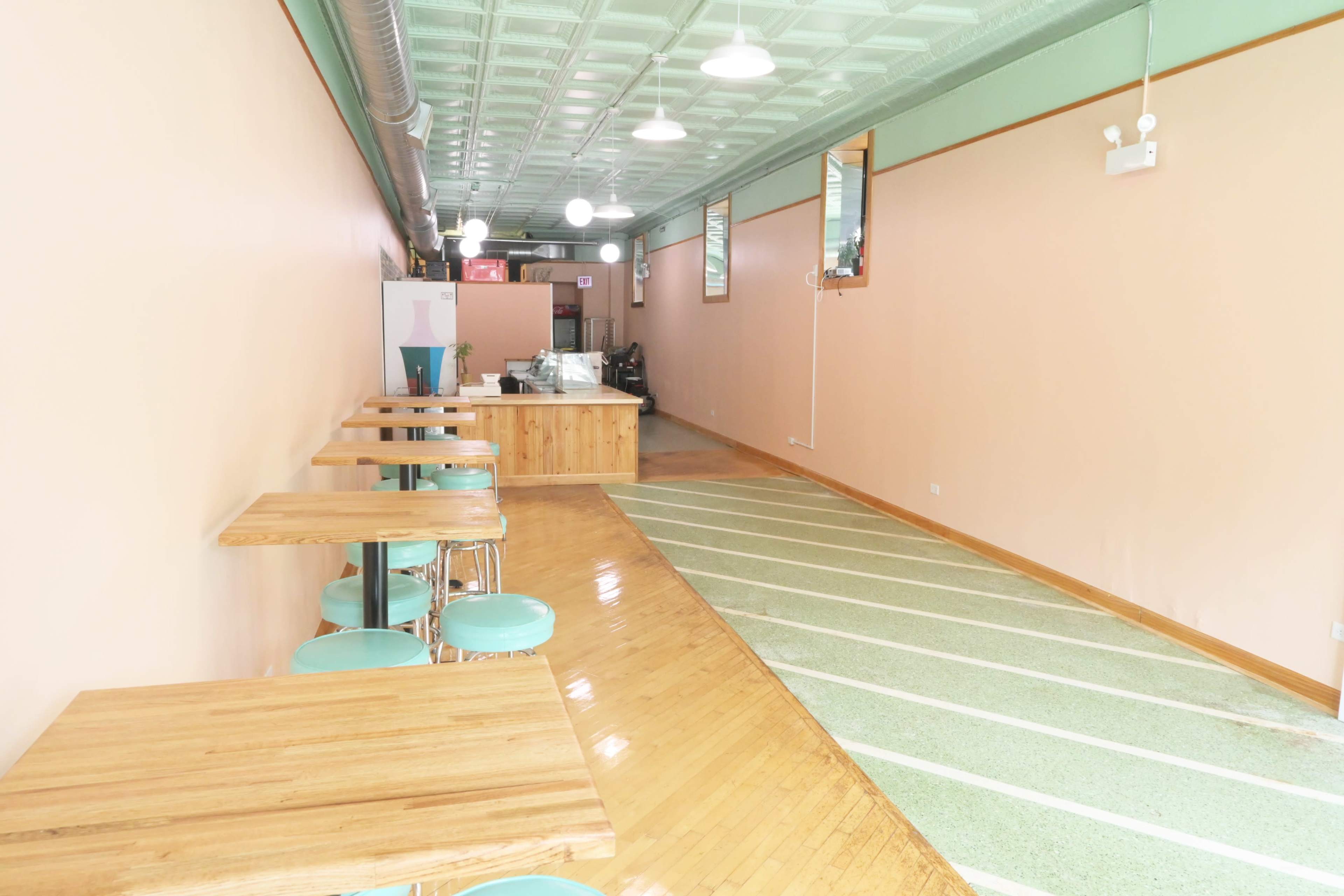 A narrow, brightly lit interior of a café featuring wooden tables with teal stools and a service counter at the end.