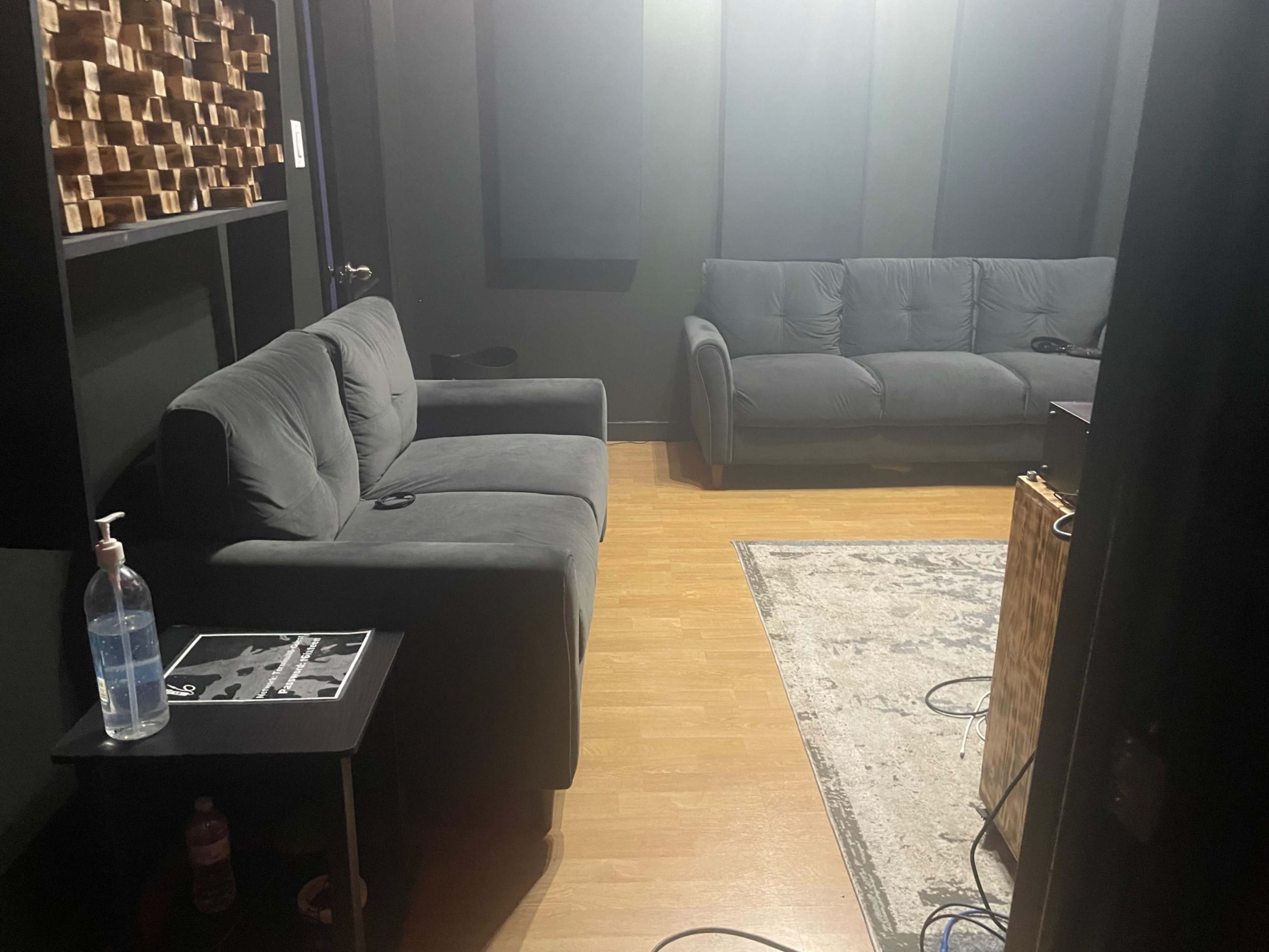 The image shows a dimly lit recording studio with a gray couch, a small table with a bottle of hand sanitizer, and a wooden wall structure in the background.