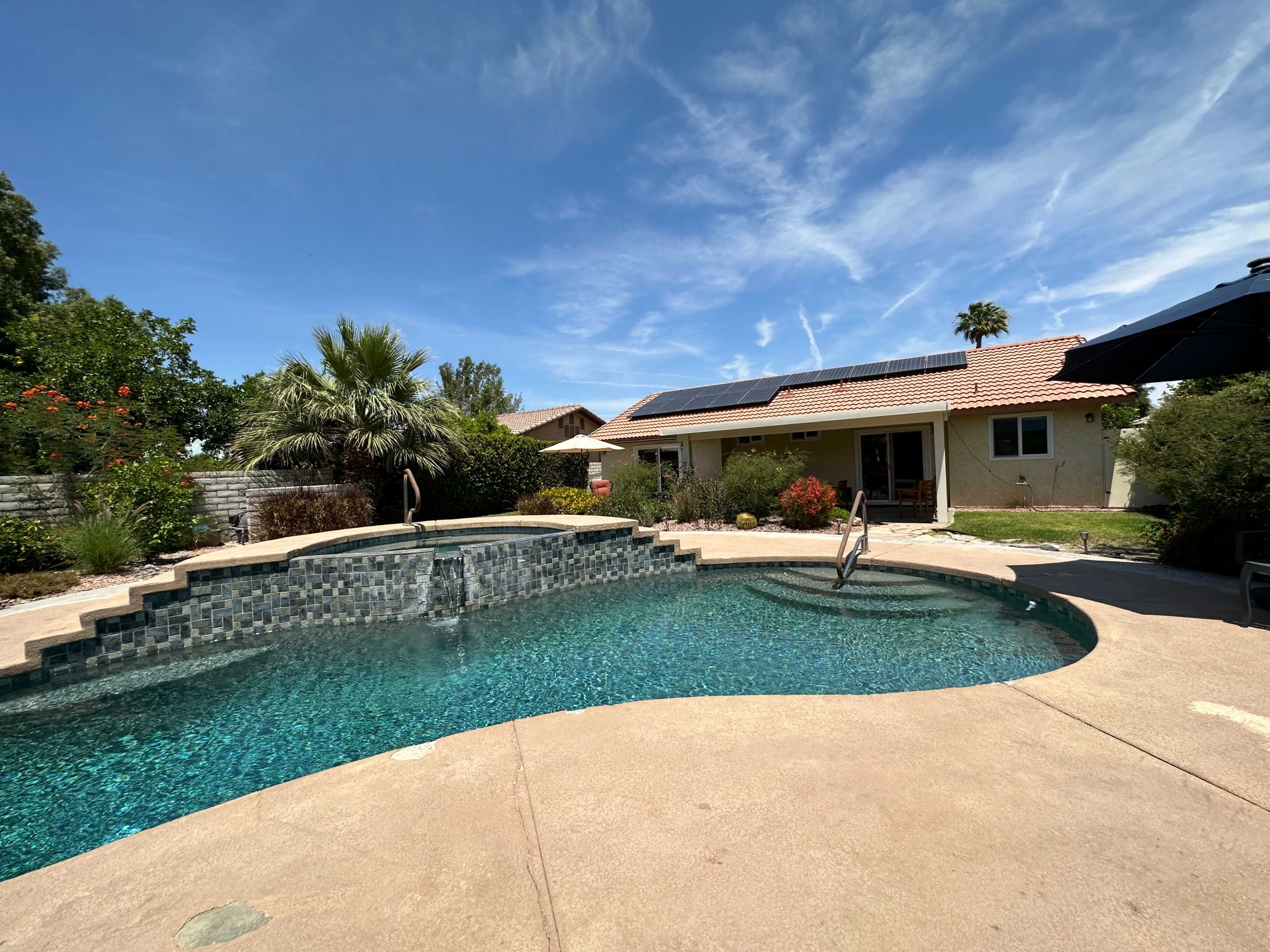 A backyard features a swimming pool with a spa, surrounded by tropical plants and a single-story house with solar panels on the roof.