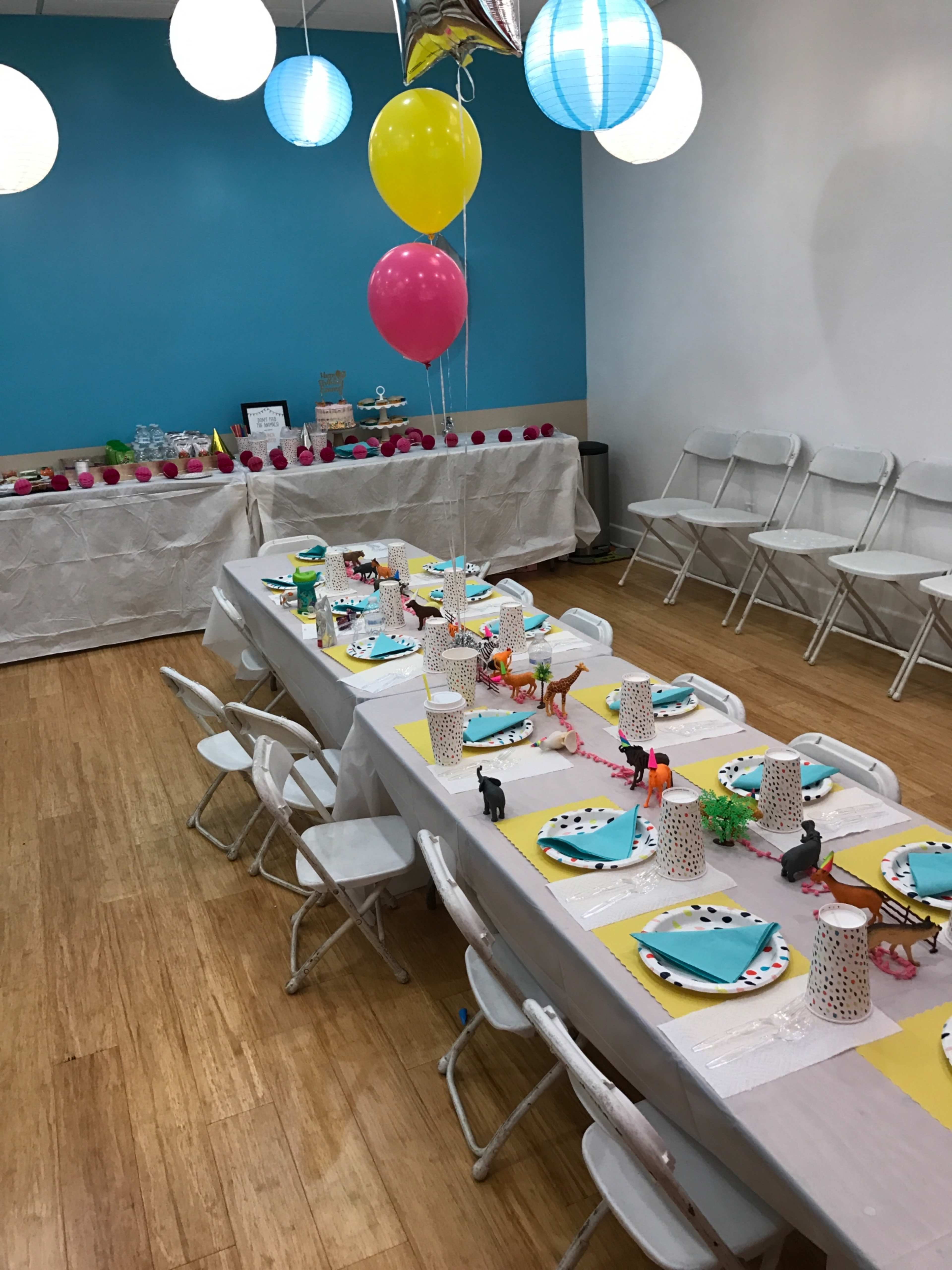 A decorated party room features a long table set with colorful plates, animal-themed centerpieces, and balloons, surrounded by white folding chairs.