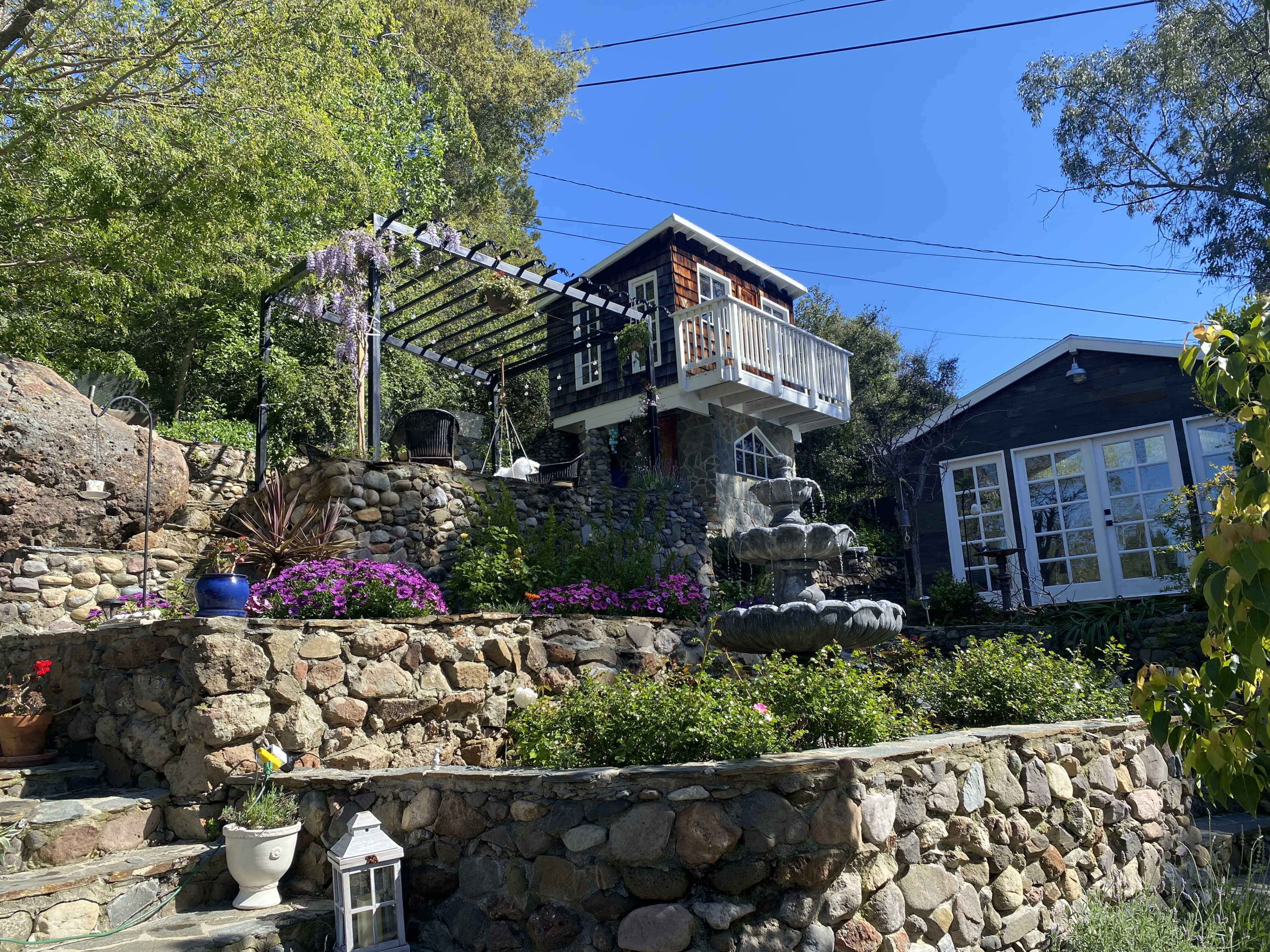A landscaped hillside features a stone terrace with a cascading water fountain and two distinct structures surrounded by various plants and flowers.