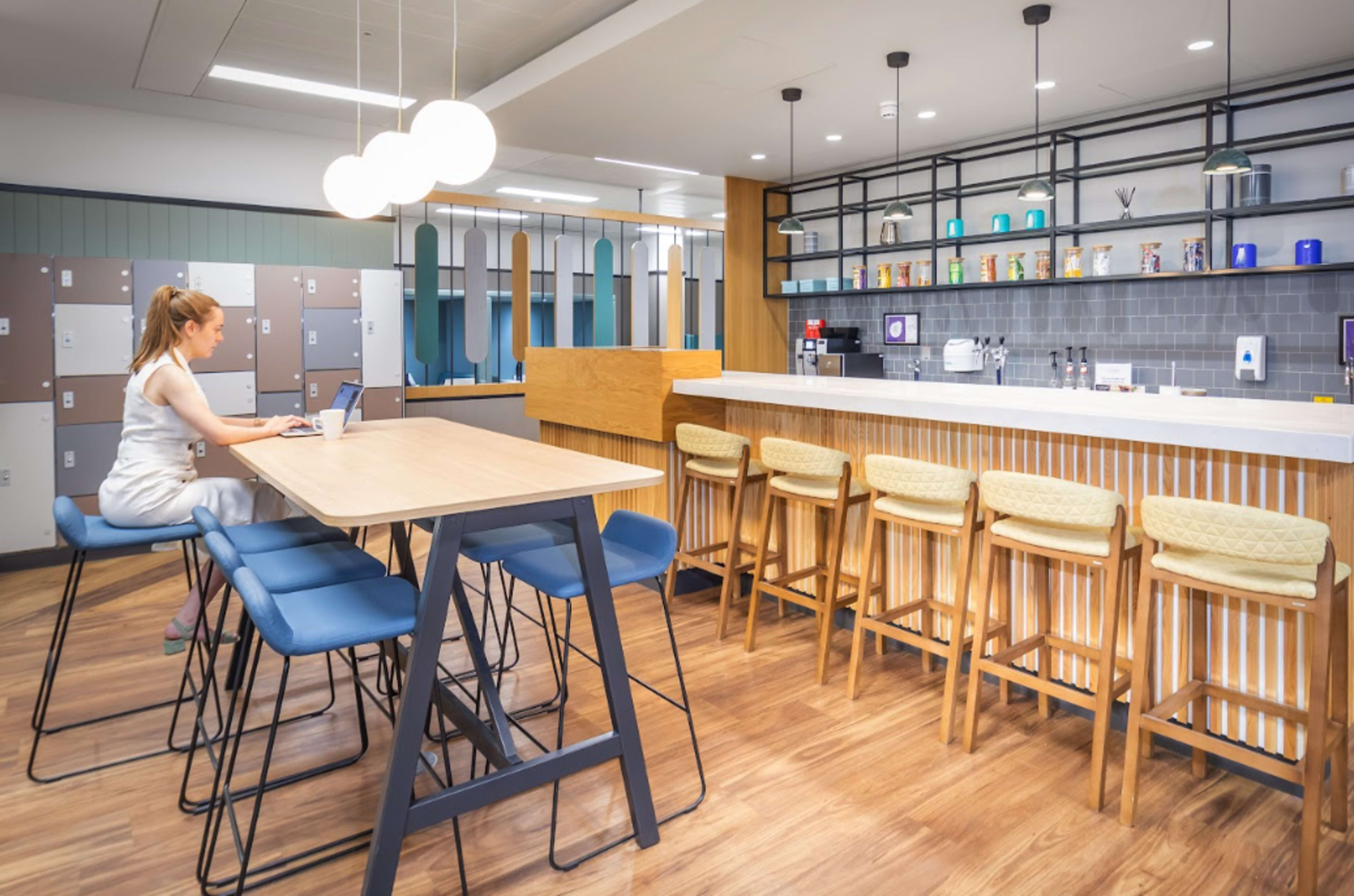 A woman is seated at a table using a laptop in a modern kitchen area featuring bar stools and lockers in the background.