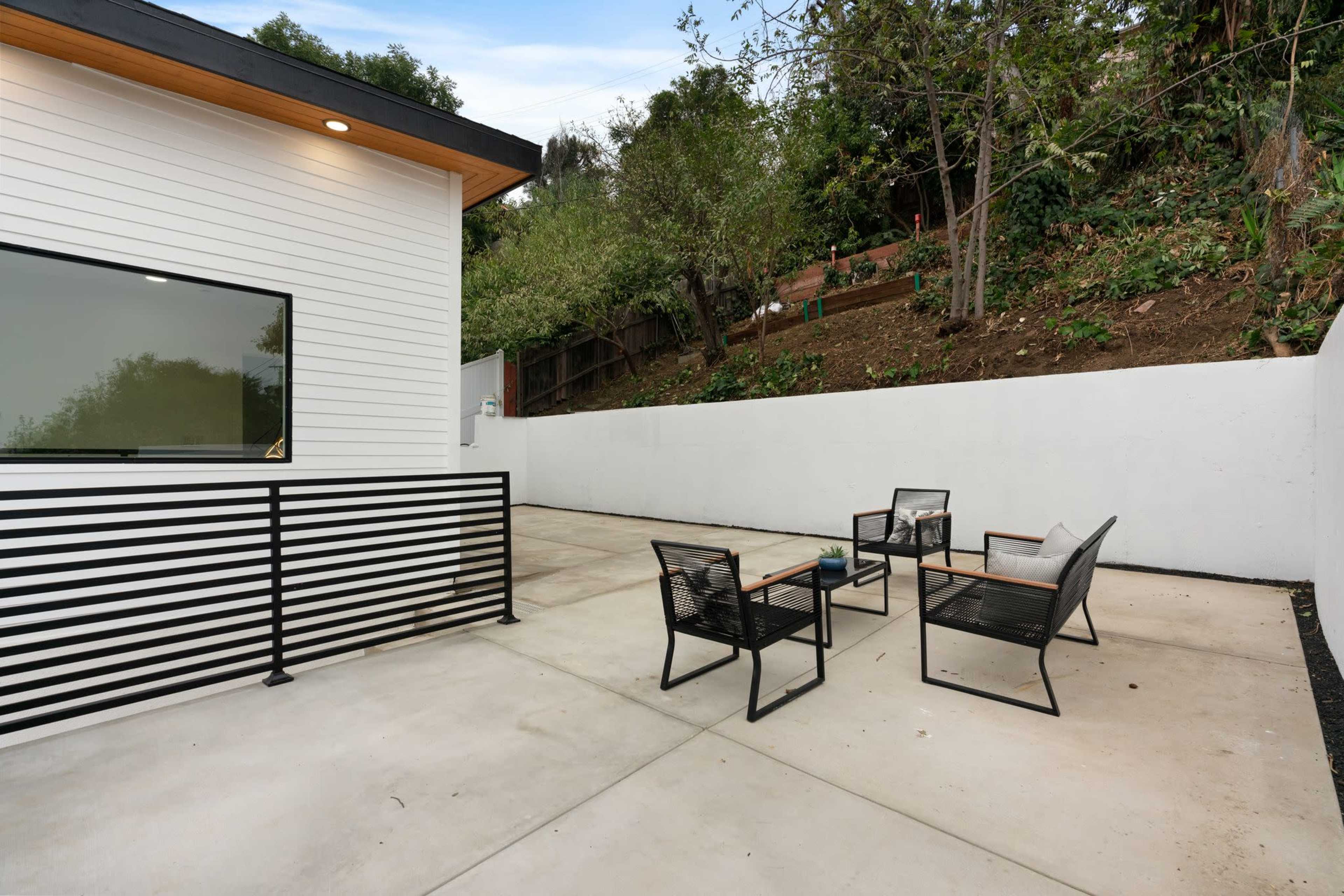 A modern patio area features four black chairs arranged around a small table, adjacent to a white wall and greenery on a hillside.