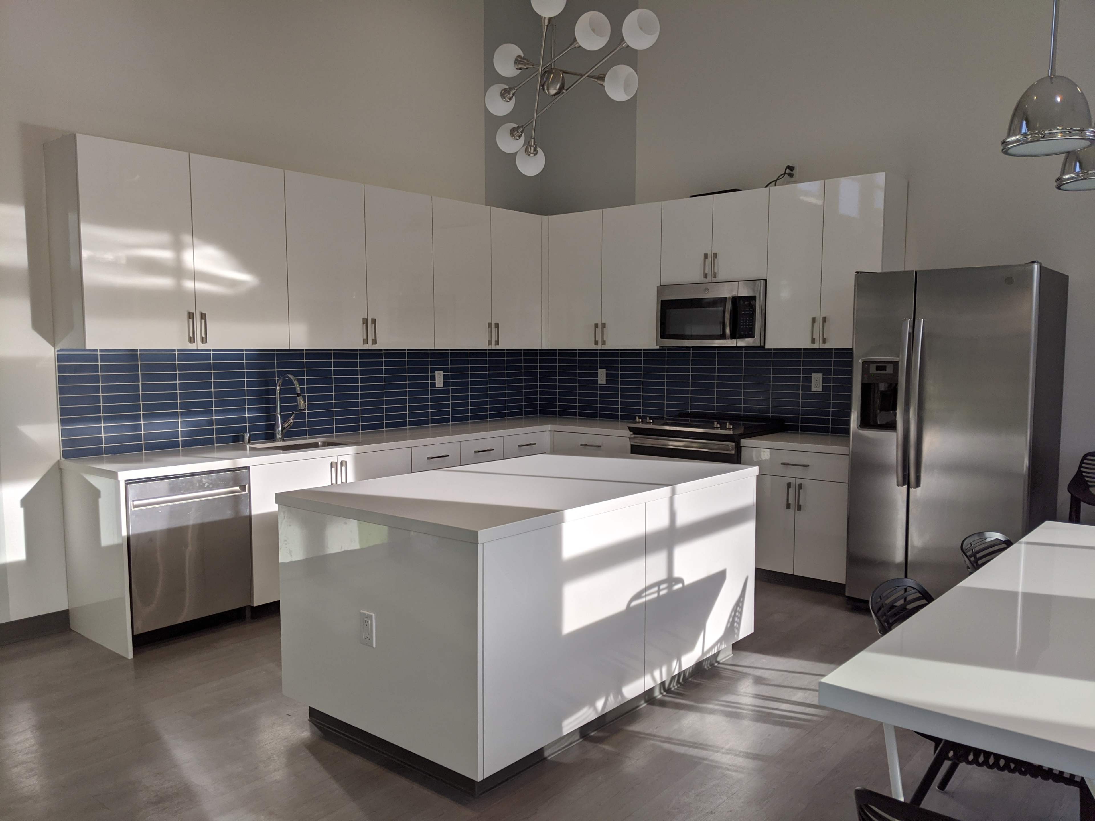 The image shows a modern kitchen featuring white cabinetry, a blue tiled backsplash, stainless steel appliances, and a central island with a light countertop.