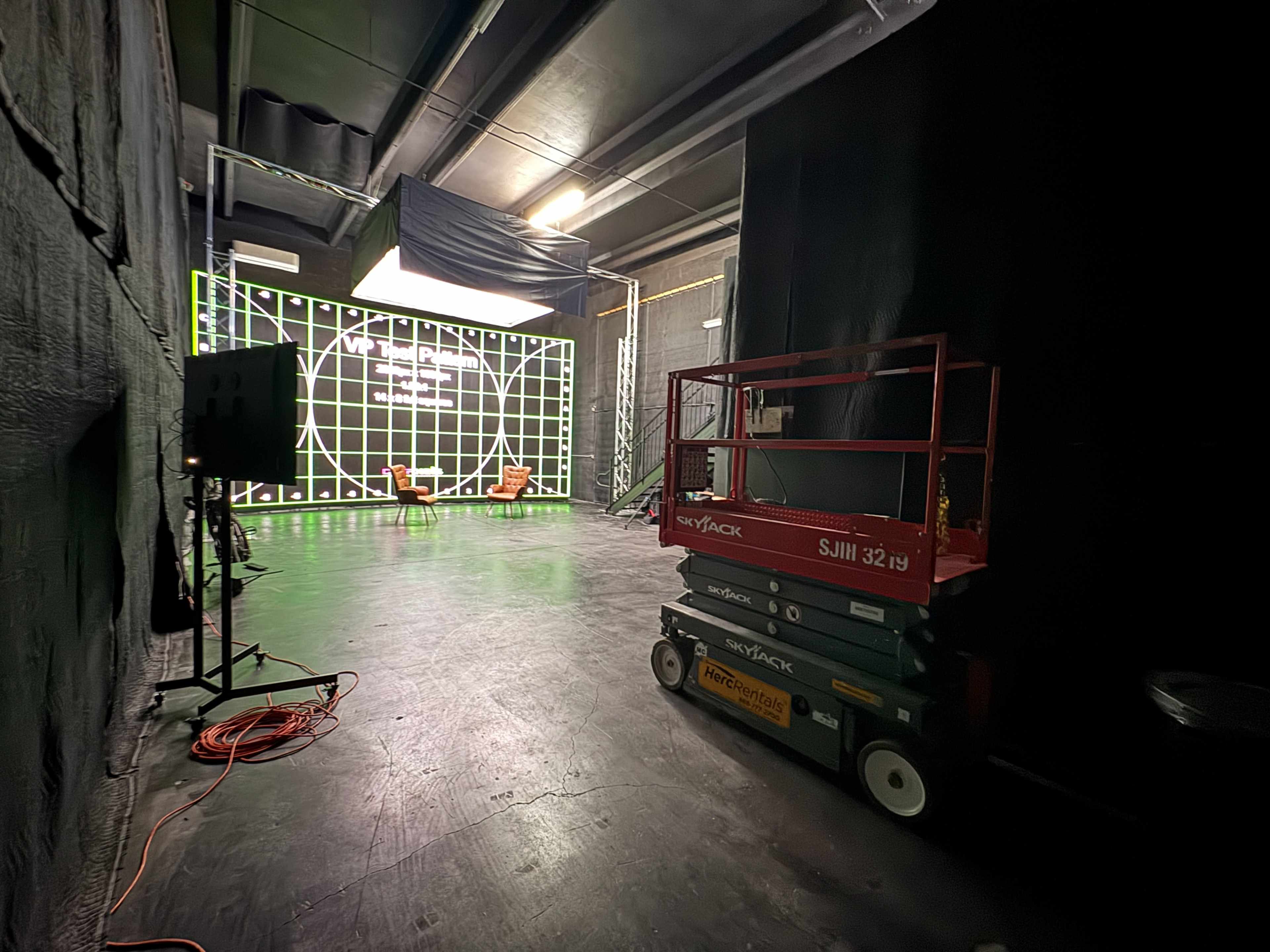 The image shows a dimly lit recording studio with a grid-patterned backdrop, two chairs, and a lift placed near the foreground.