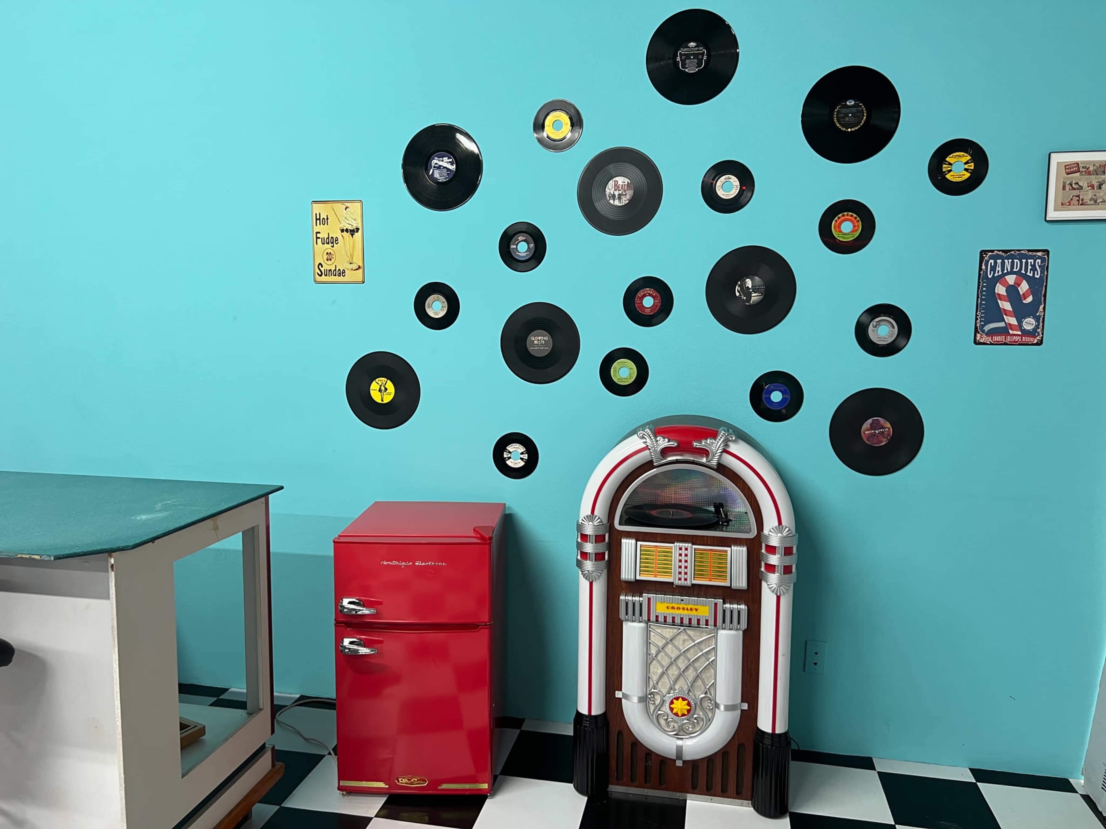 The image shows a turquoise wall decorated with black vinyl records, alongside a red refrigerator and a jukebox.