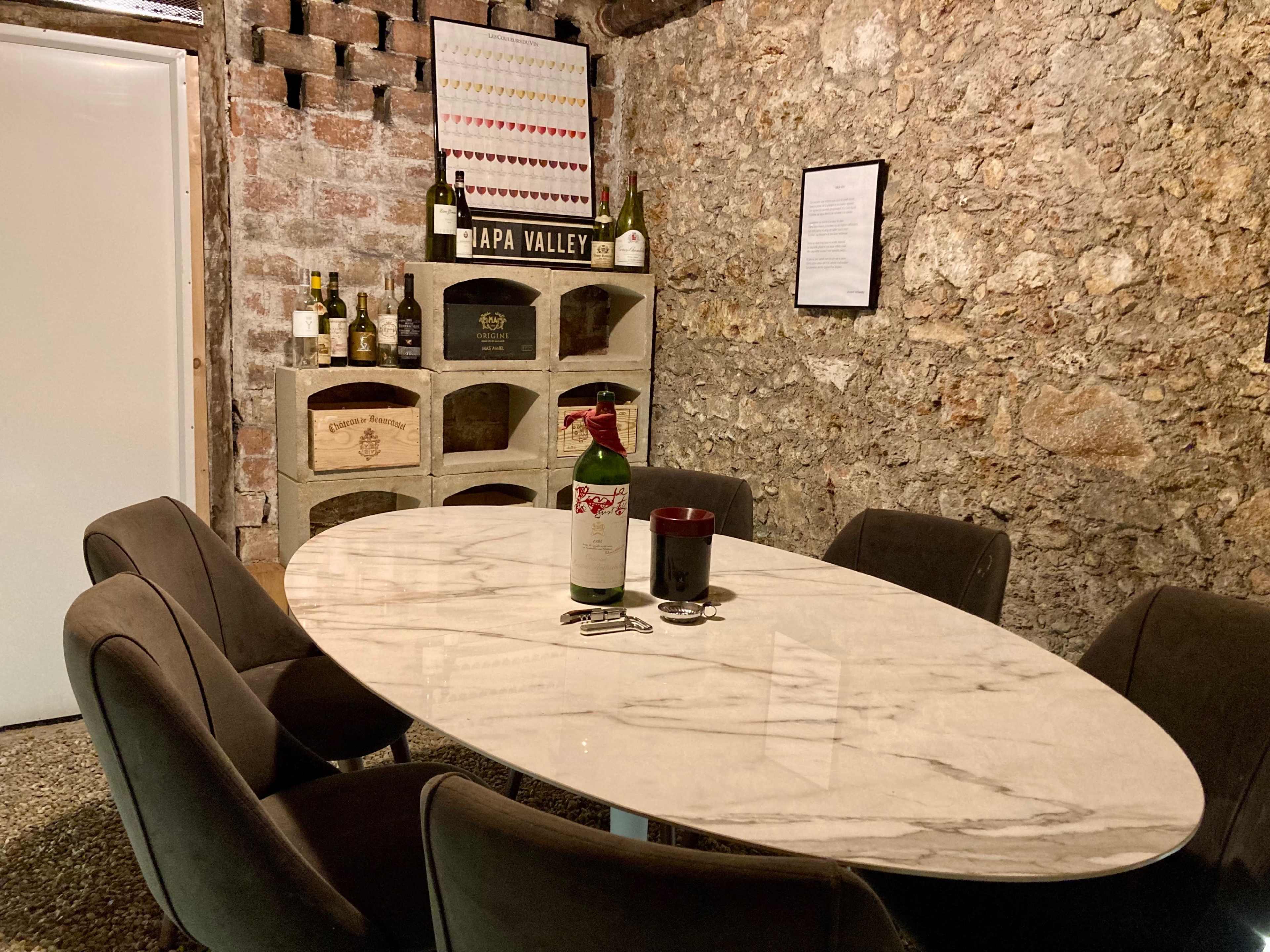 A marble-topped table with six upholstered chairs is set in a rustic stone cellar, surrounded by wine bottles and wine storage racks.