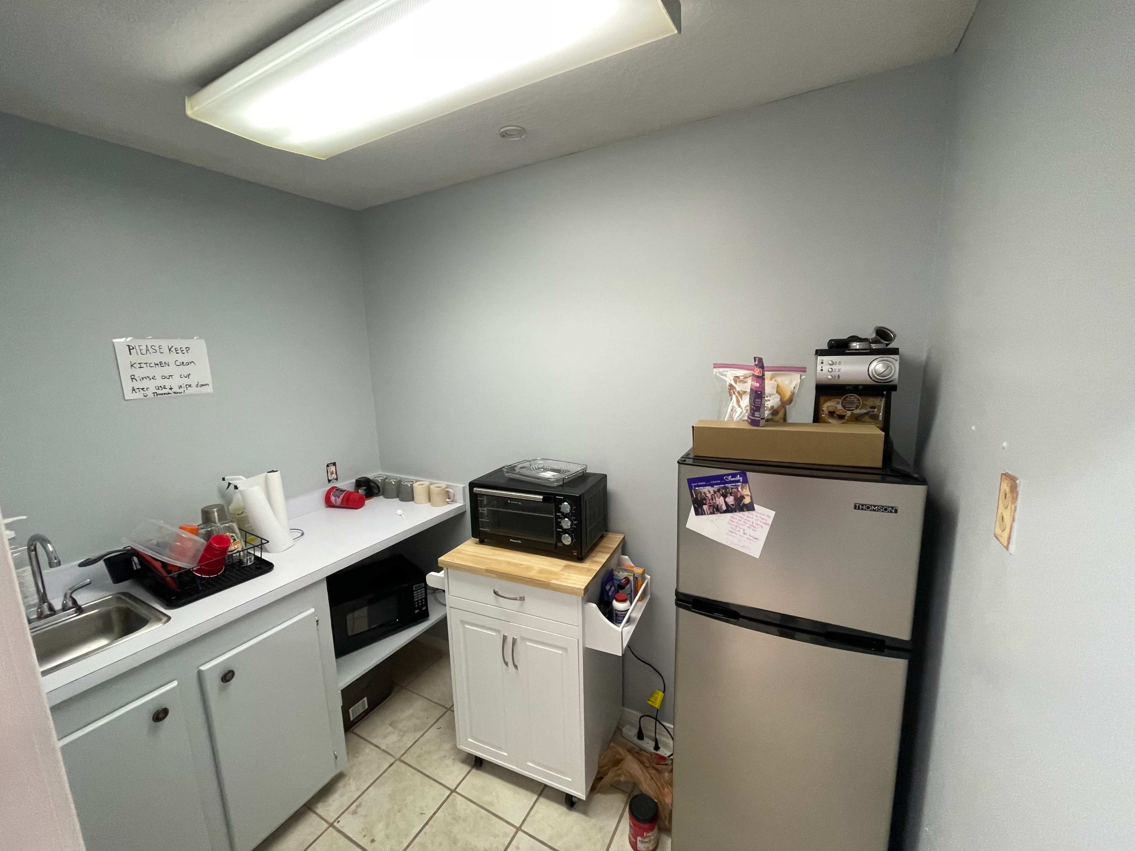The image shows a small kitchen area with a sink, countertop, a refrigerator, and a microwave on top of a cabinet.