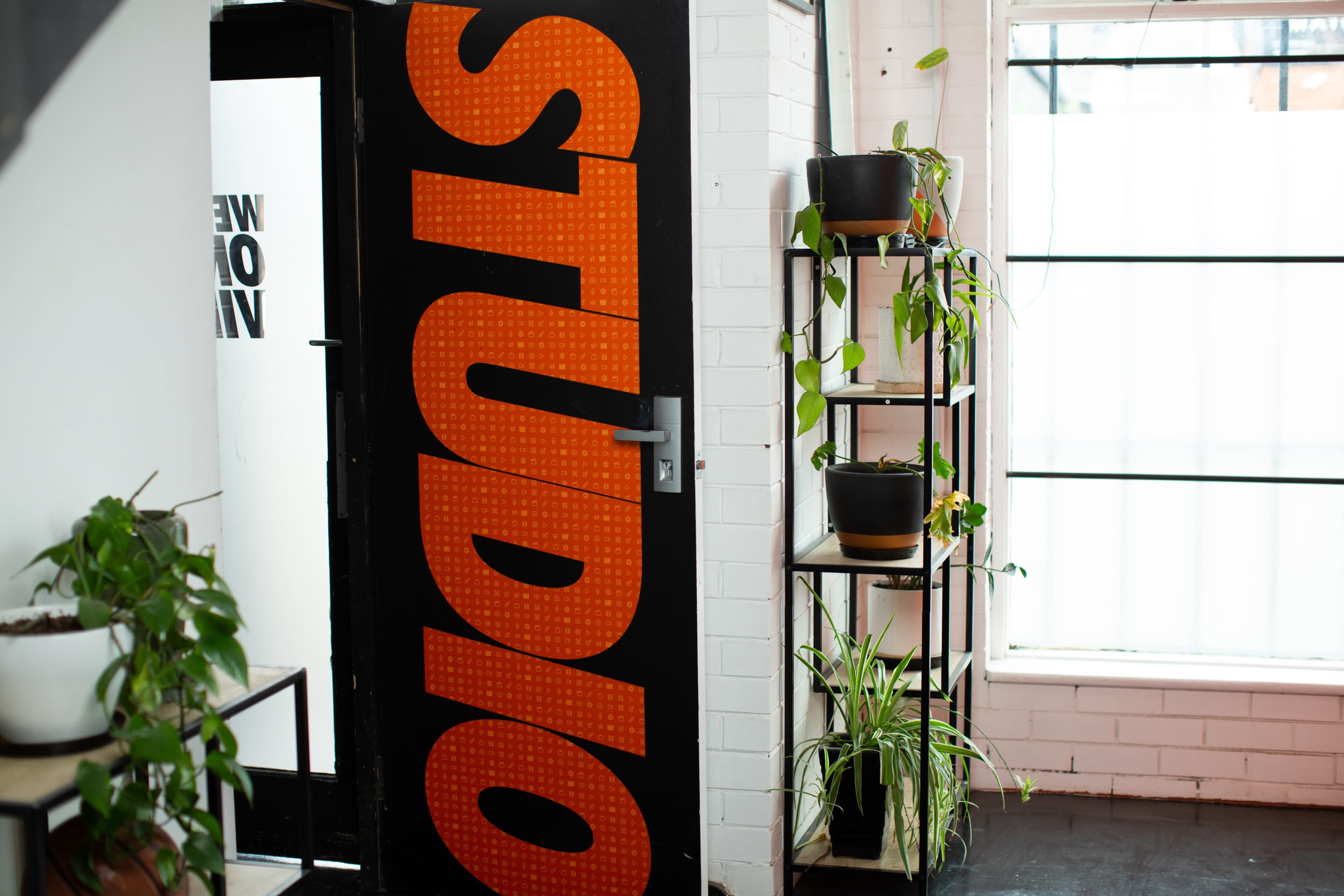 A black door with the word "STUDIO" in orange lettering opens into a room with potted plants on a shelf and natural light from a large window.