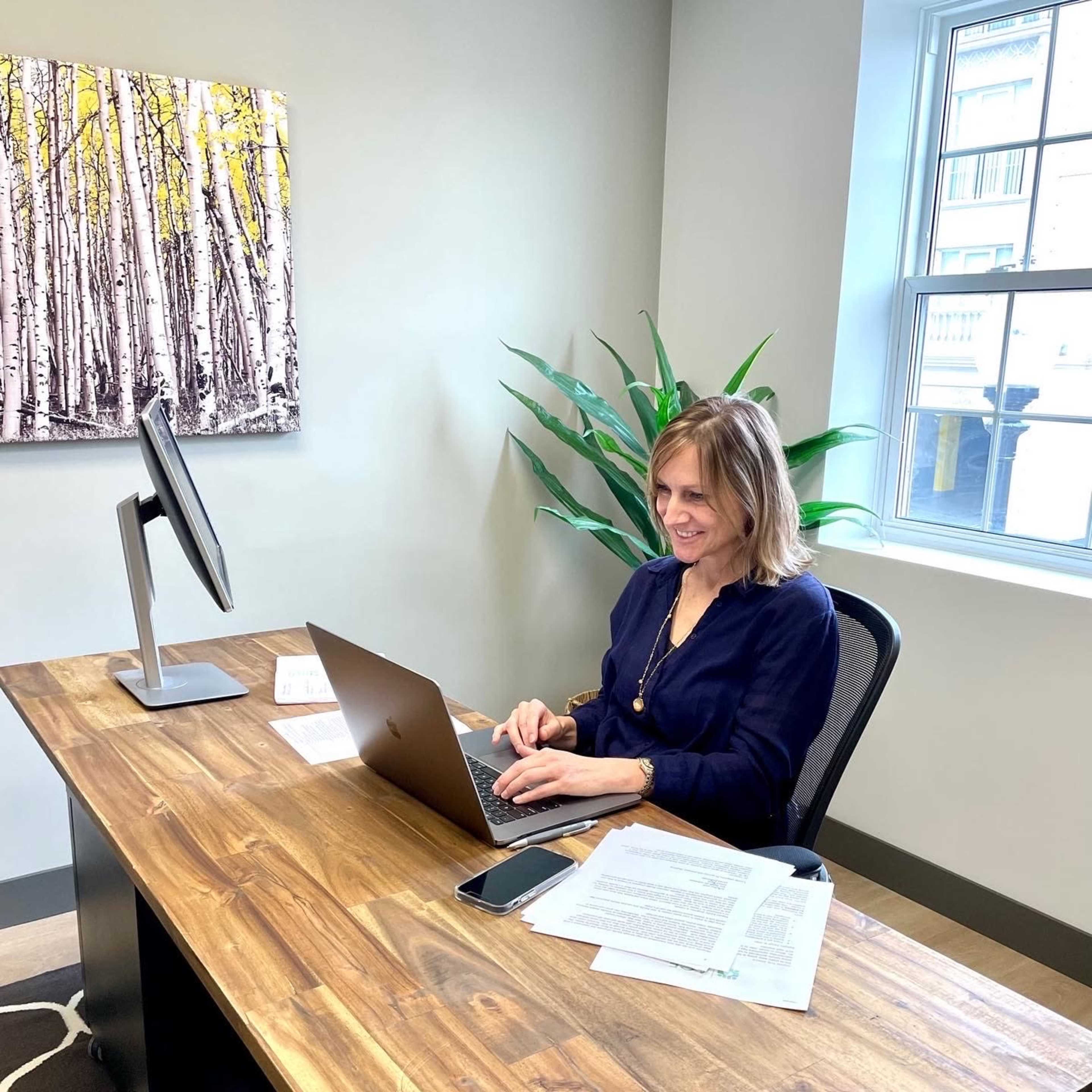 A woman sits at a wooden desk, working on a laptop with a smartphone and documents nearby, in an office setting.