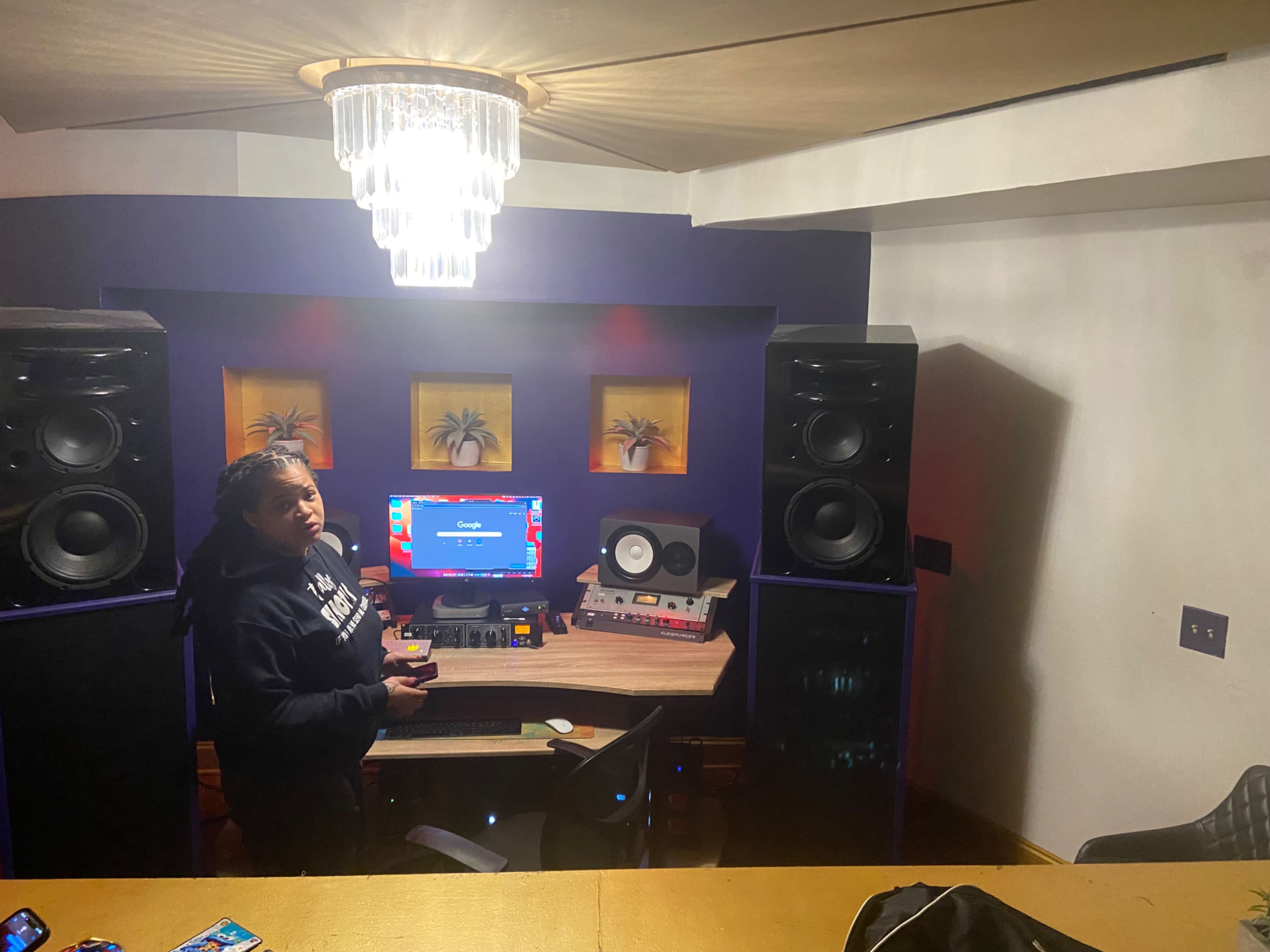 A person stands in a home recording studio equipped with a computer, audio equipment, and large speakers.