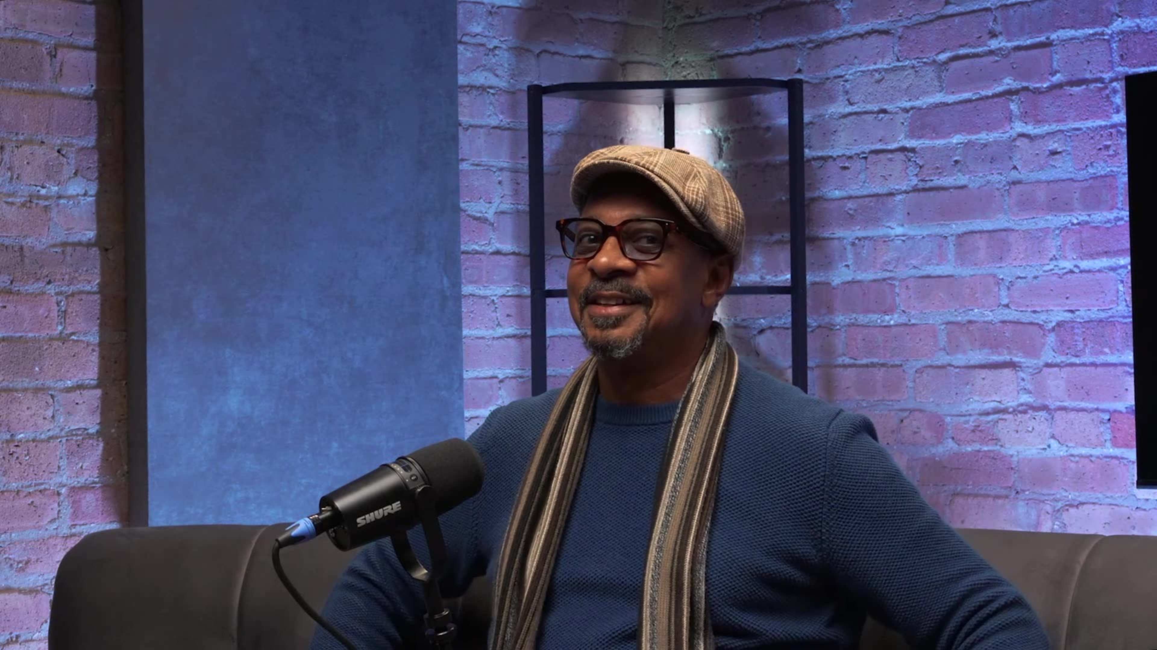 A man wearing glasses and a cap sits in front of a microphone in a well-lit room with brick walls.