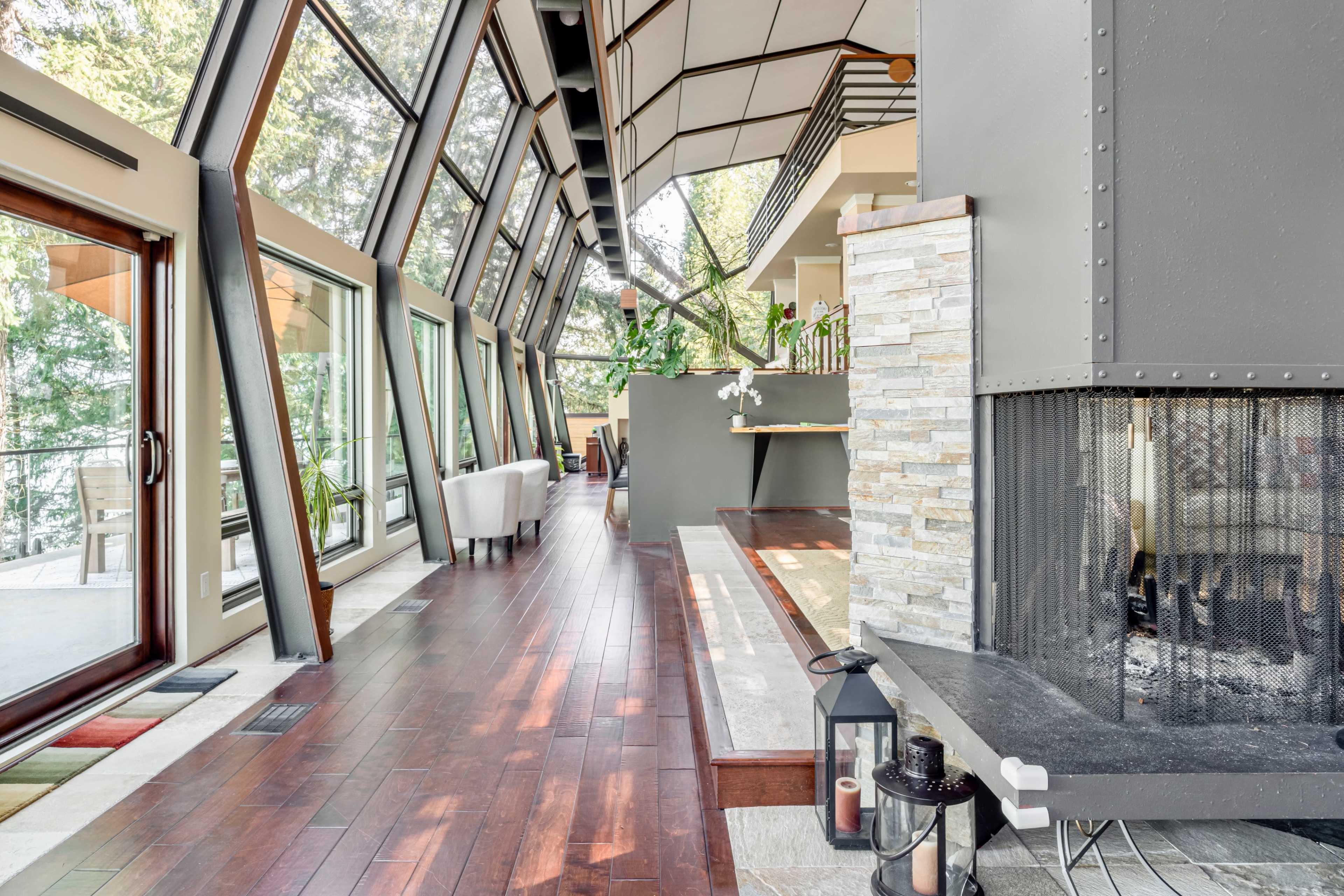 A modern interior space with large windows, wooden flooring, and a stone fireplace, featuring a view of greenery outside.