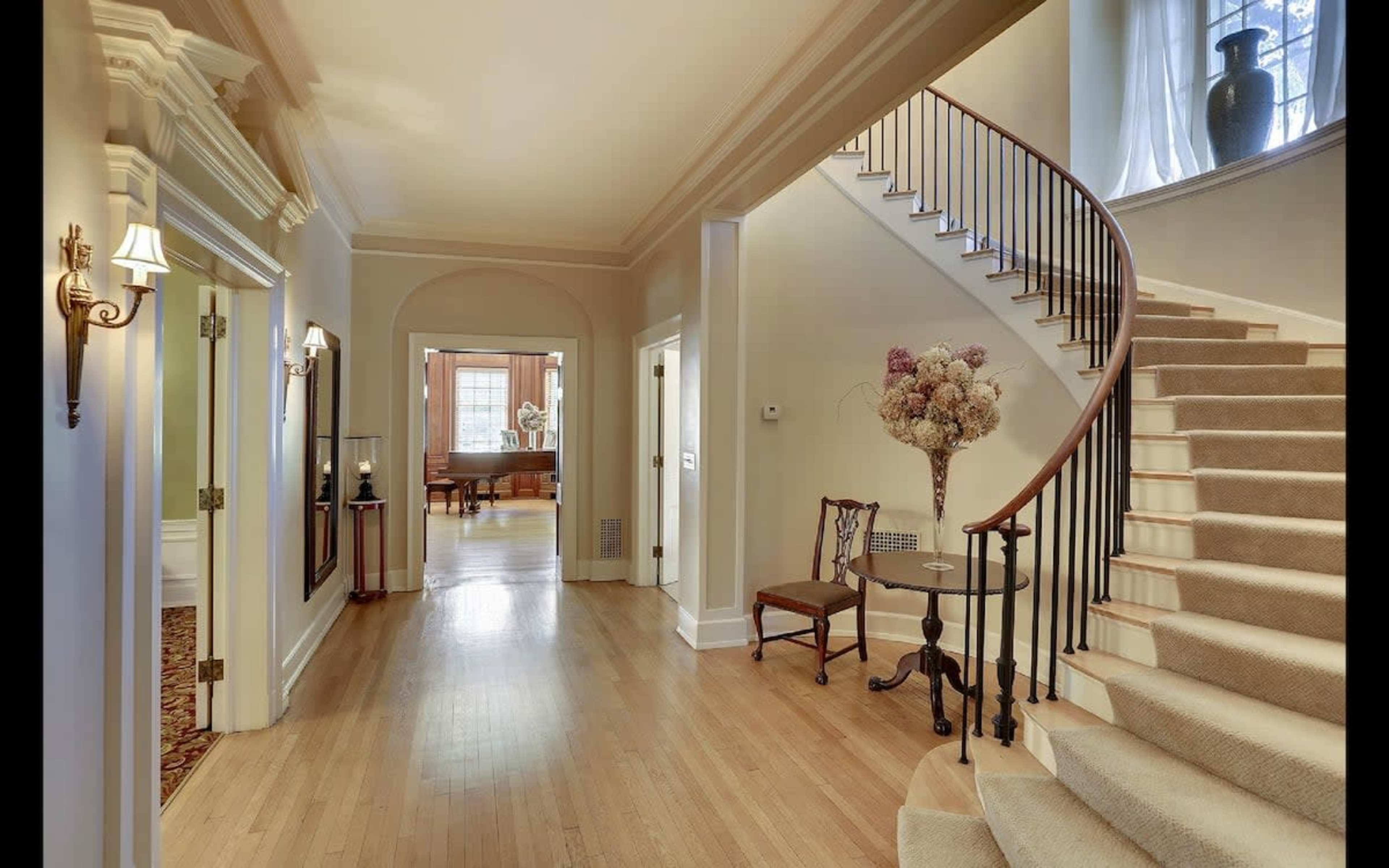 A spacious hallway with a staircase on the right, leading to an upper level, and a floral arrangement on a table near the entrance.