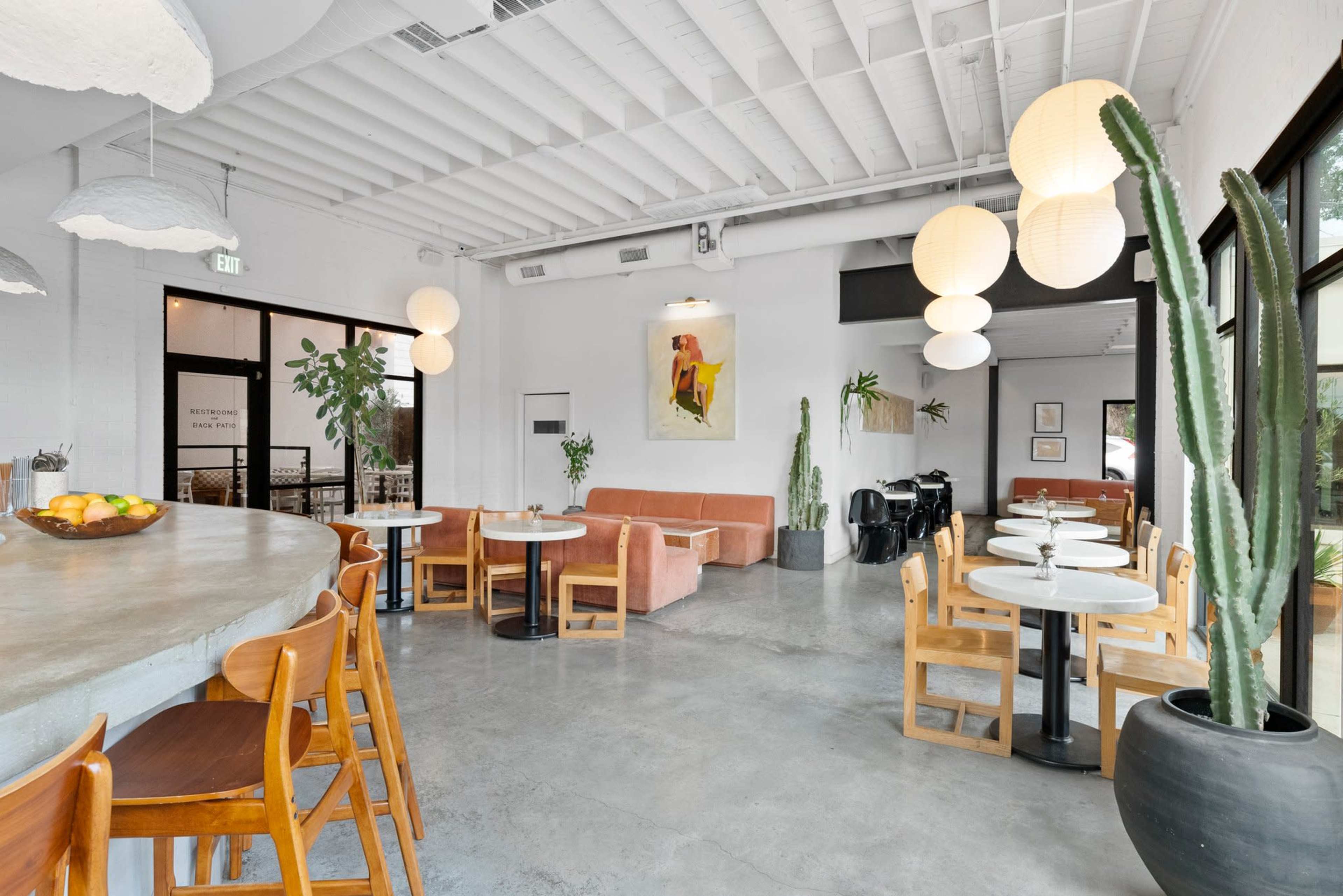 A modern café features a concrete floor, pastel seating, a large bar area, and decorative plants.