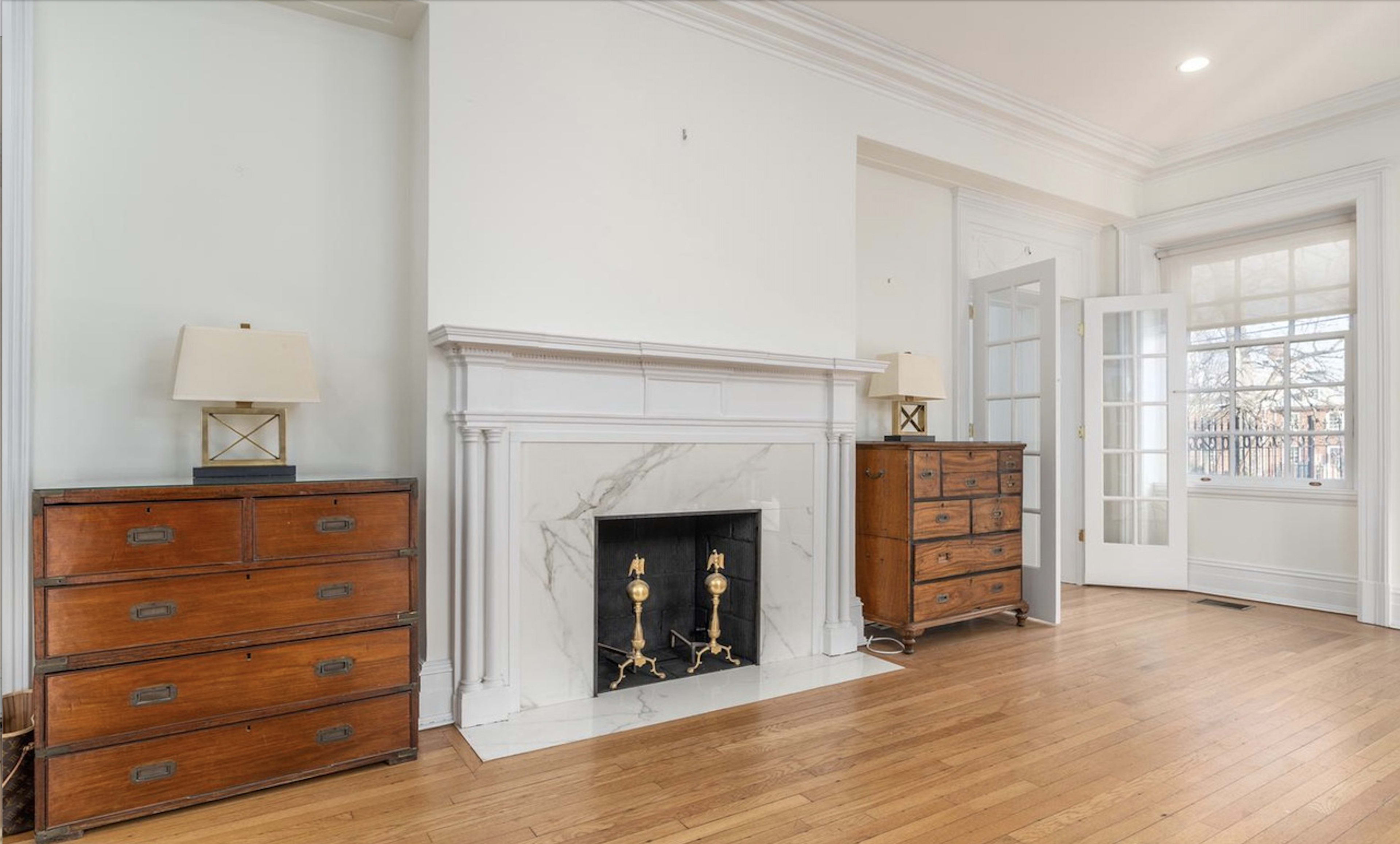 A living room with a marble fireplace centered between two wooden dressers and a set of French doors leading to a balcony.