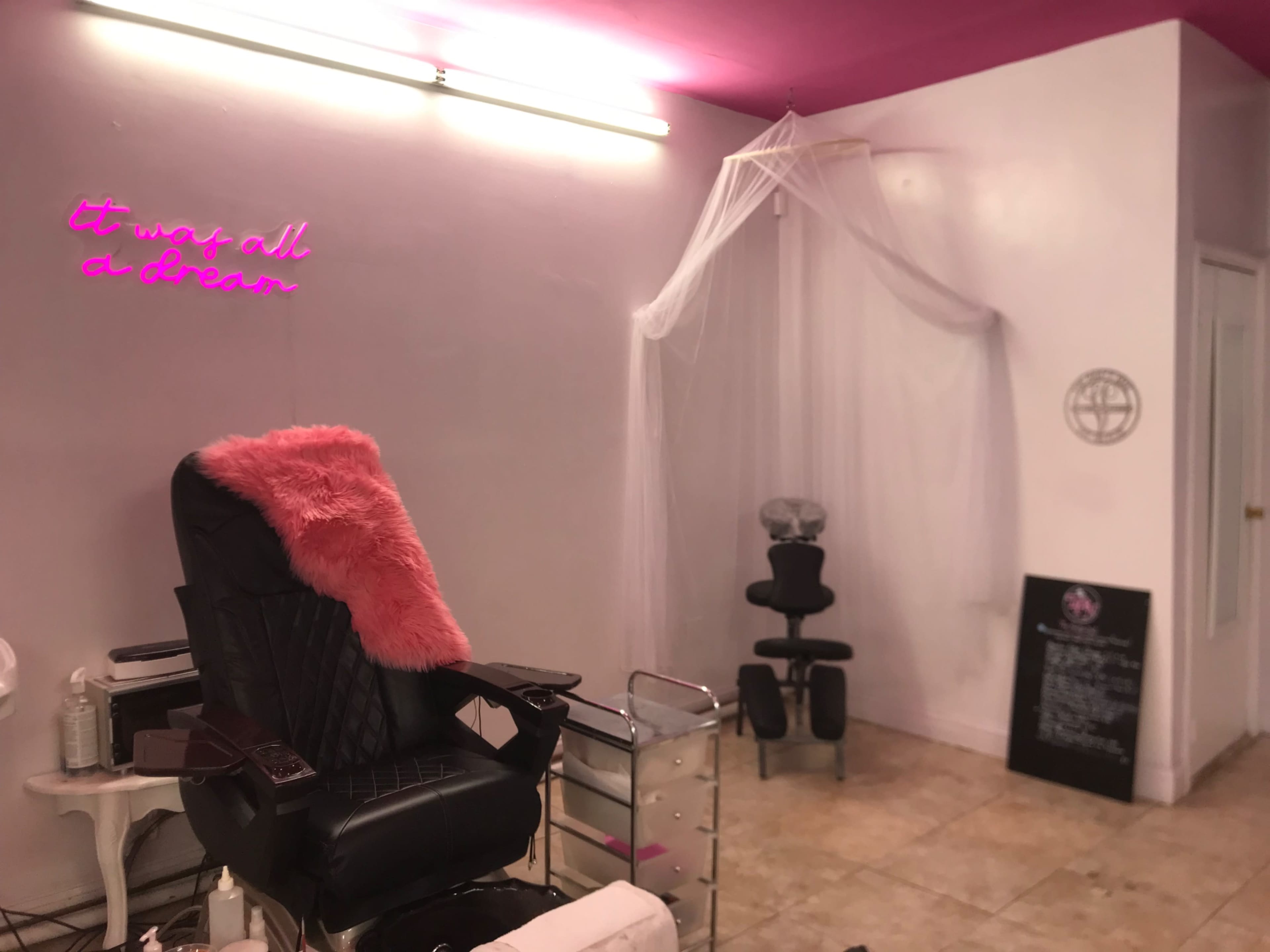 The interior of a beauty salon features a black salon chair with a pink faux fur throw, a draped area in the corner, and a neon sign that says "It was all a dream."