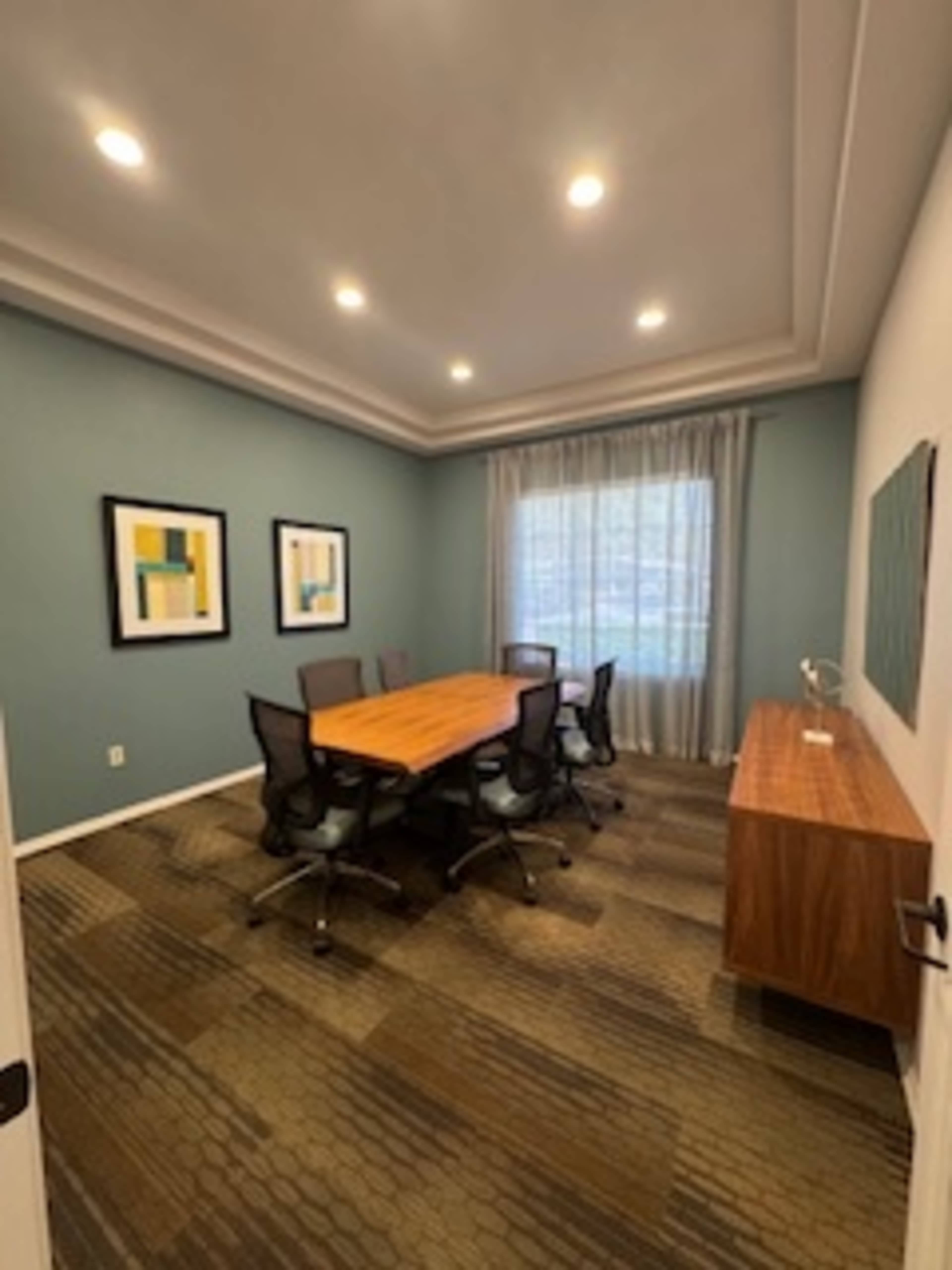 A conference room with a wooden table surrounded by six chairs, framed artwork on the walls, and a window with sheer curtains.