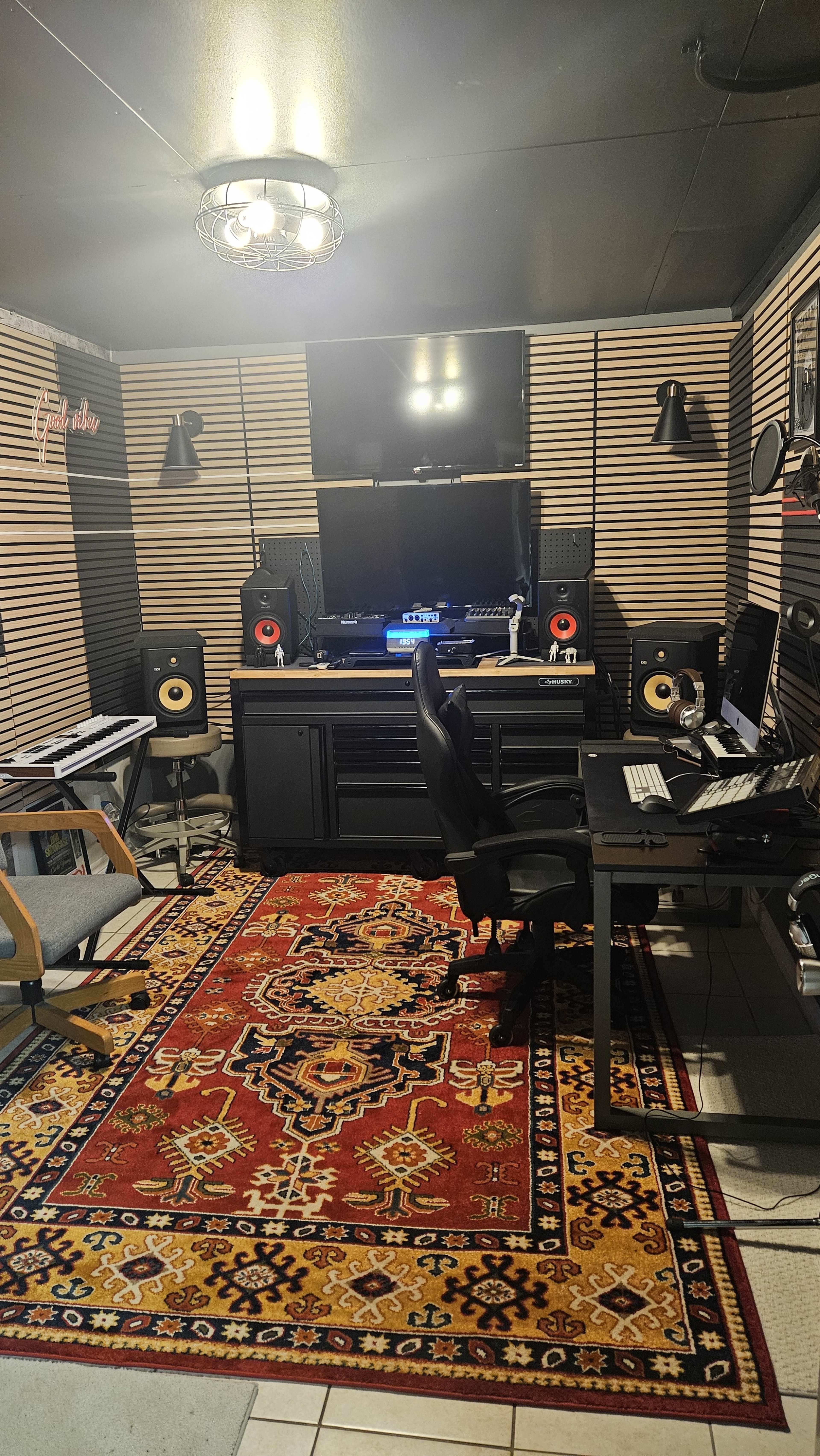 The image depicts a music studio with a large wall-mounted TV, sound speakers, and two desks arranged on a patterned rug.