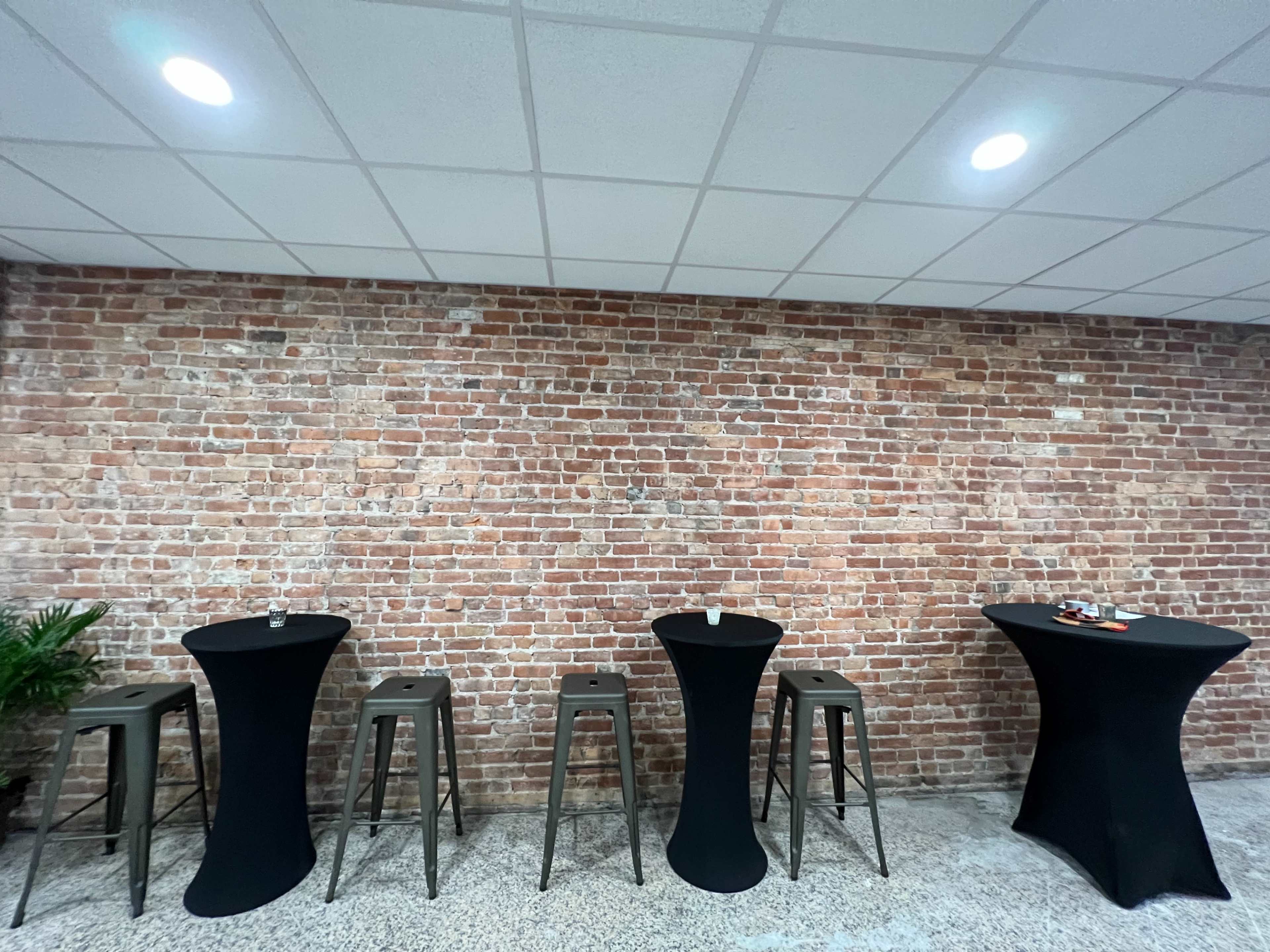 A room with a brick wall and several tall, black cocktail tables arranged in front of it.