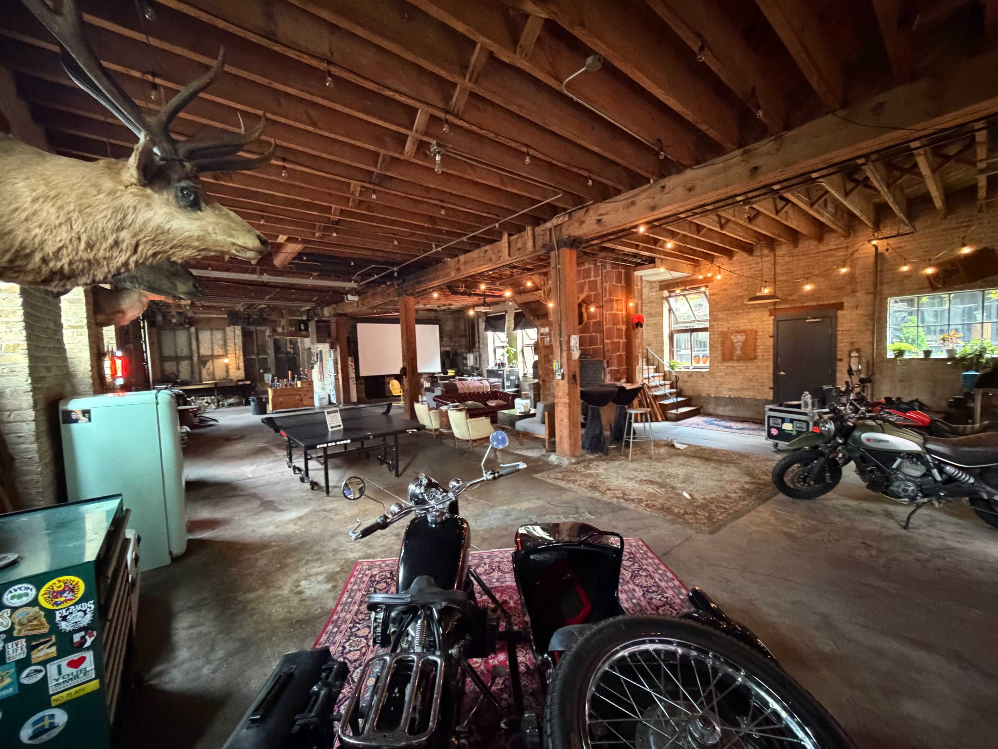 The image shows a spacious industrial-style room featuring motorcycles, a ping-pong table, and a mounted deer head, with exposed wooden beams and brick walls.