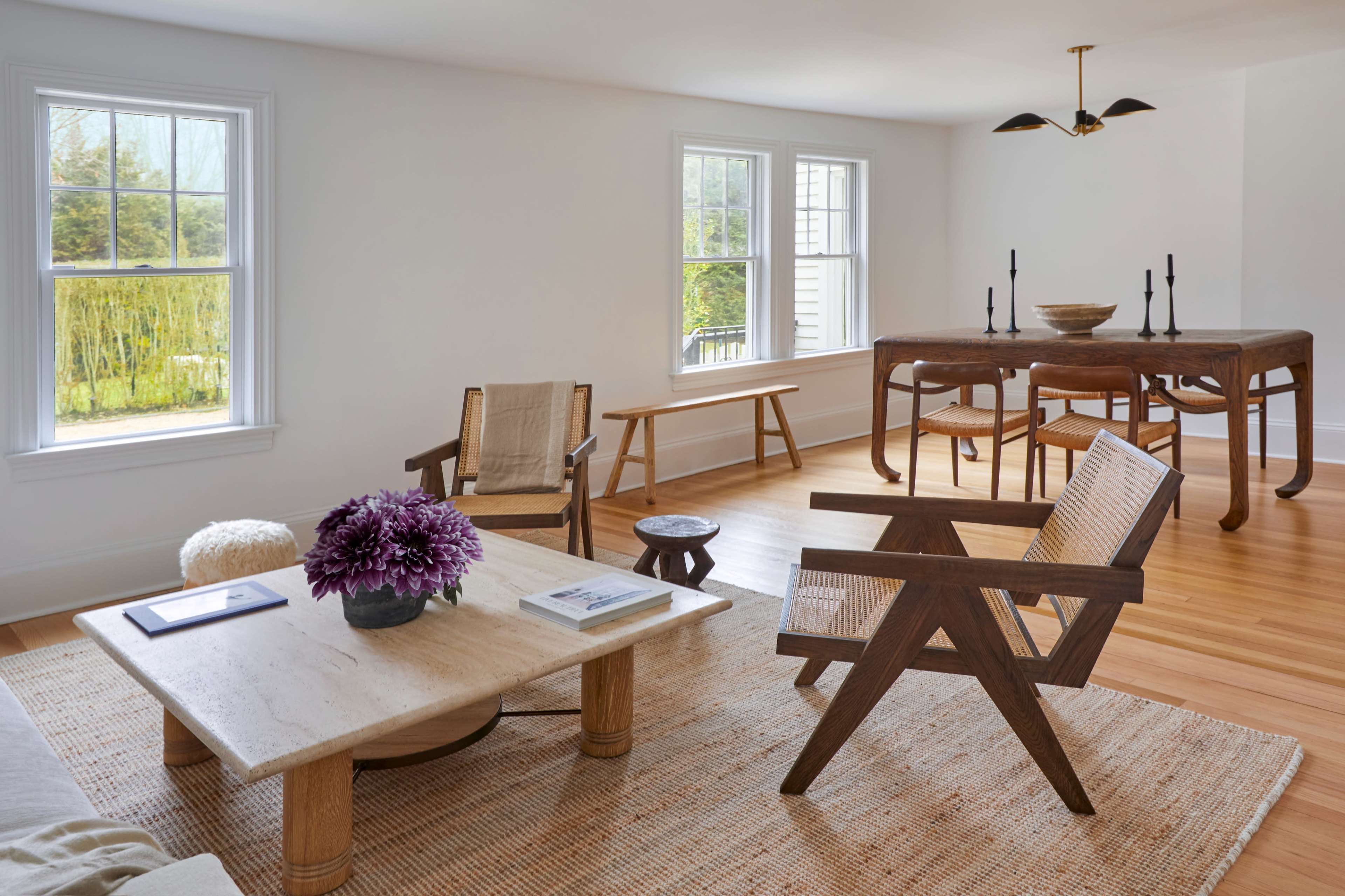 A bright, minimalist living space featuring two wooden chairs, a low coffee table, and a dining table with candles, set against a backdrop of windows and wooden flooring.