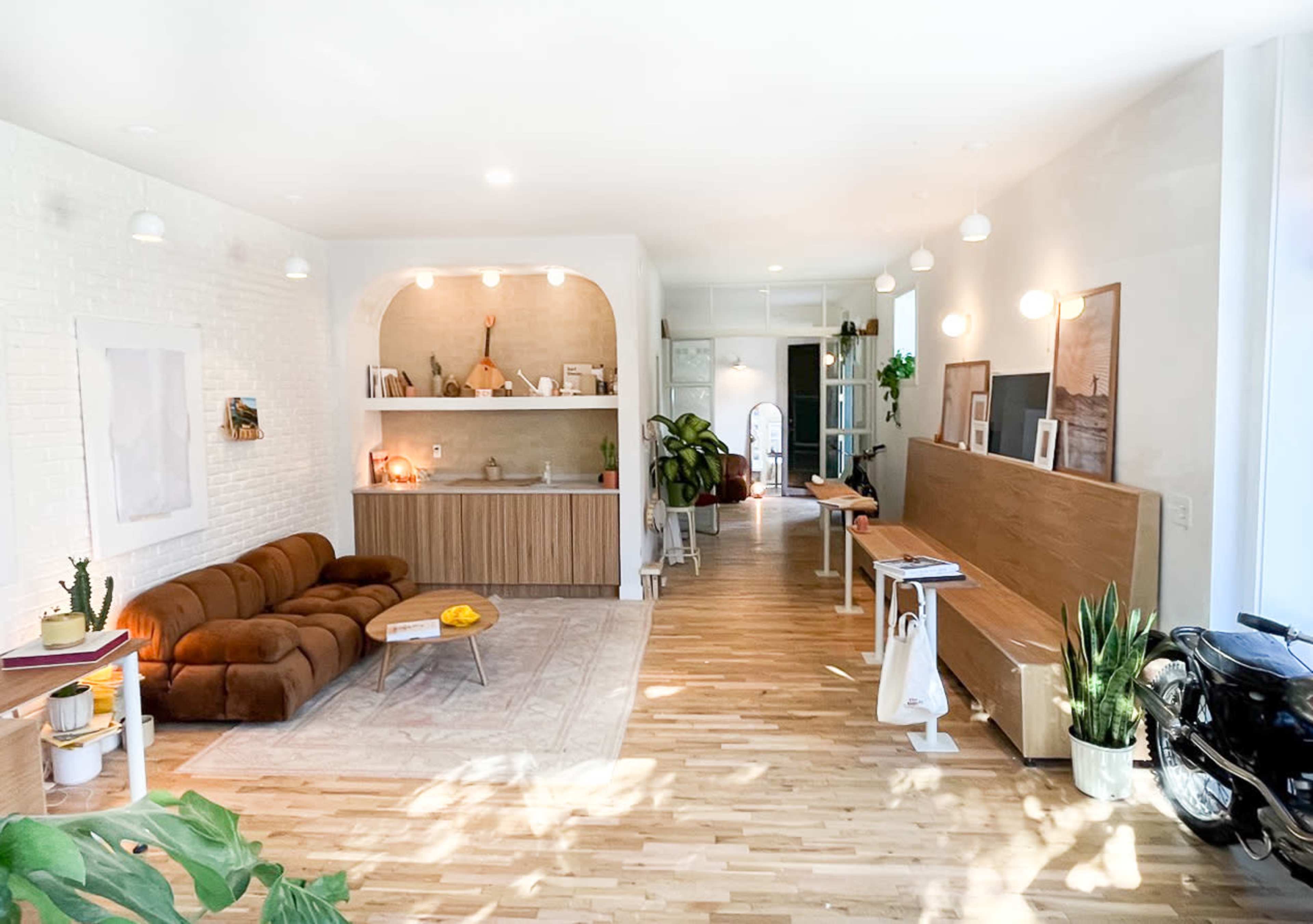 A modern, open-concept lounge area featuring a brown sofa, a light wood kitchenette, plants, and various artwork on the walls.