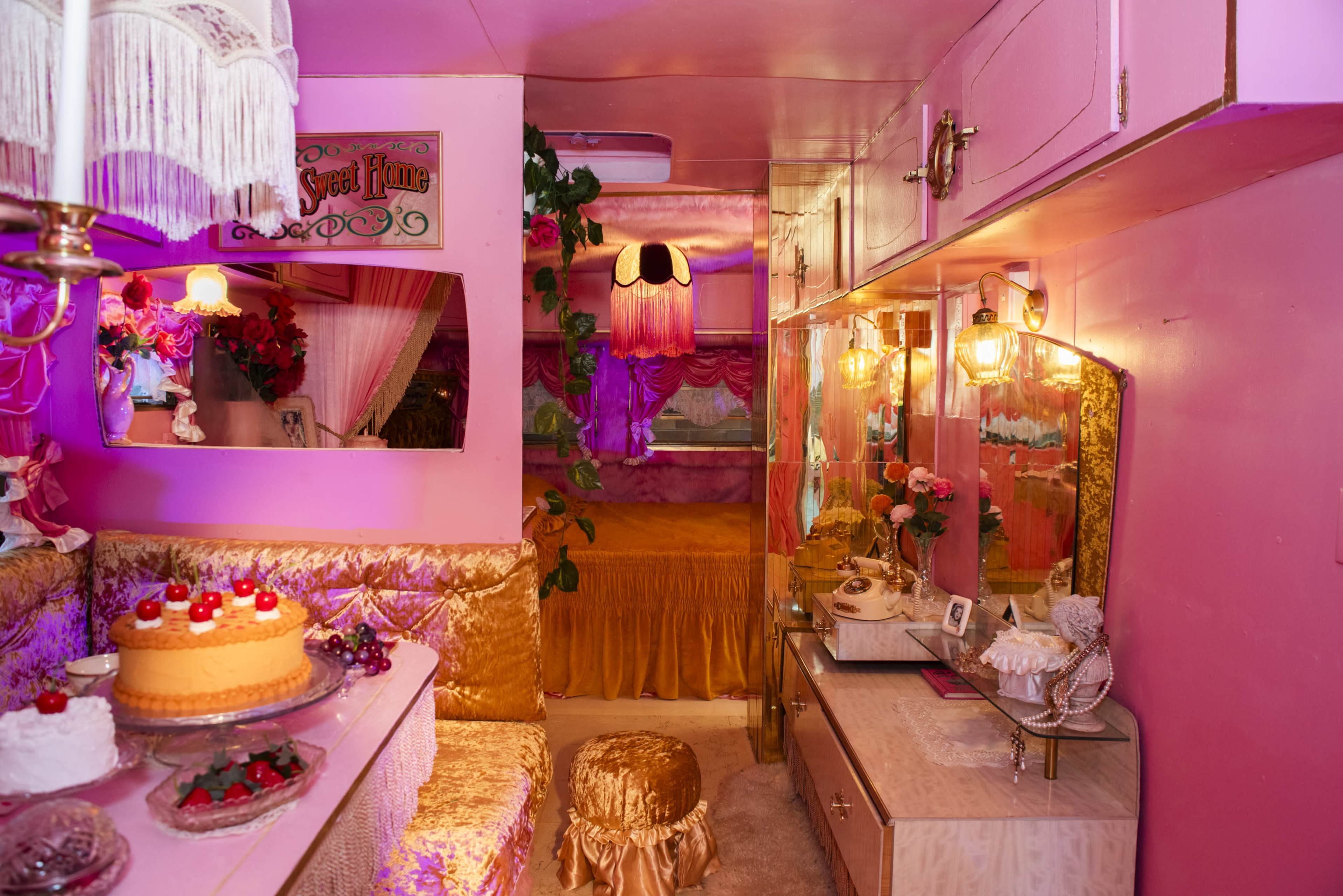 The image shows a brightly decorated pink interior of a small trailer, featuring lavish golden accents, a cozy seating area, and a mirrored cabinet adorned with flowers and desserts.