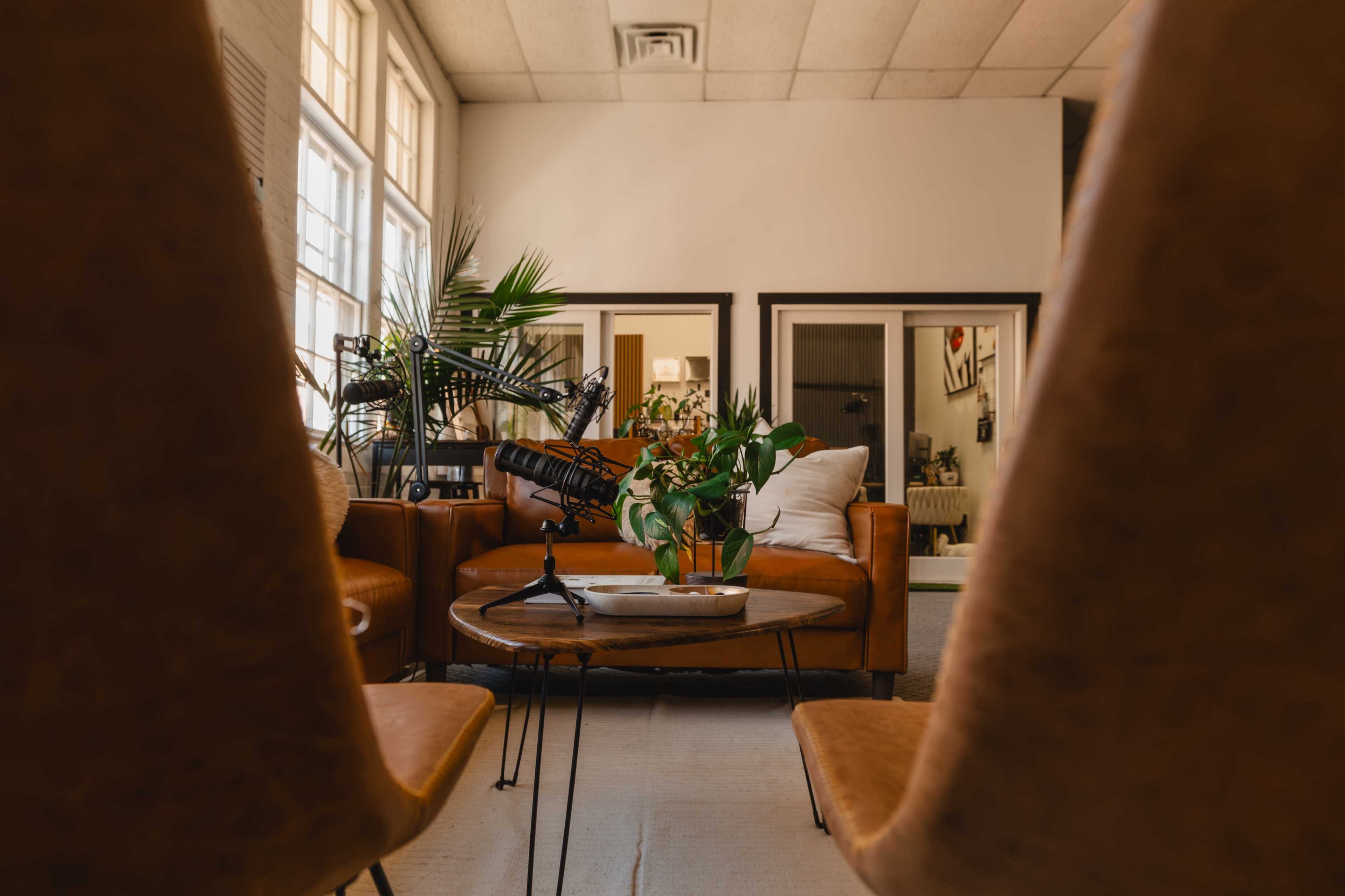 The image shows a cozy living space featuring brown leather chairs facing a circular coffee table, surrounded by plants and mirrors on the walls.