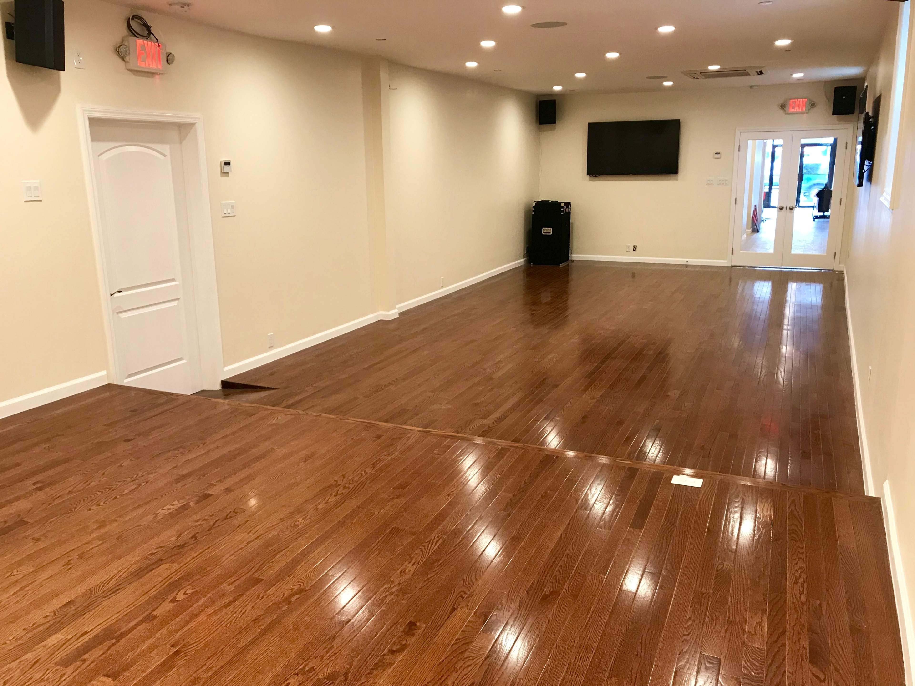A spacious, vacant interior room with hardwood floors, light walls, and several ceiling lights.