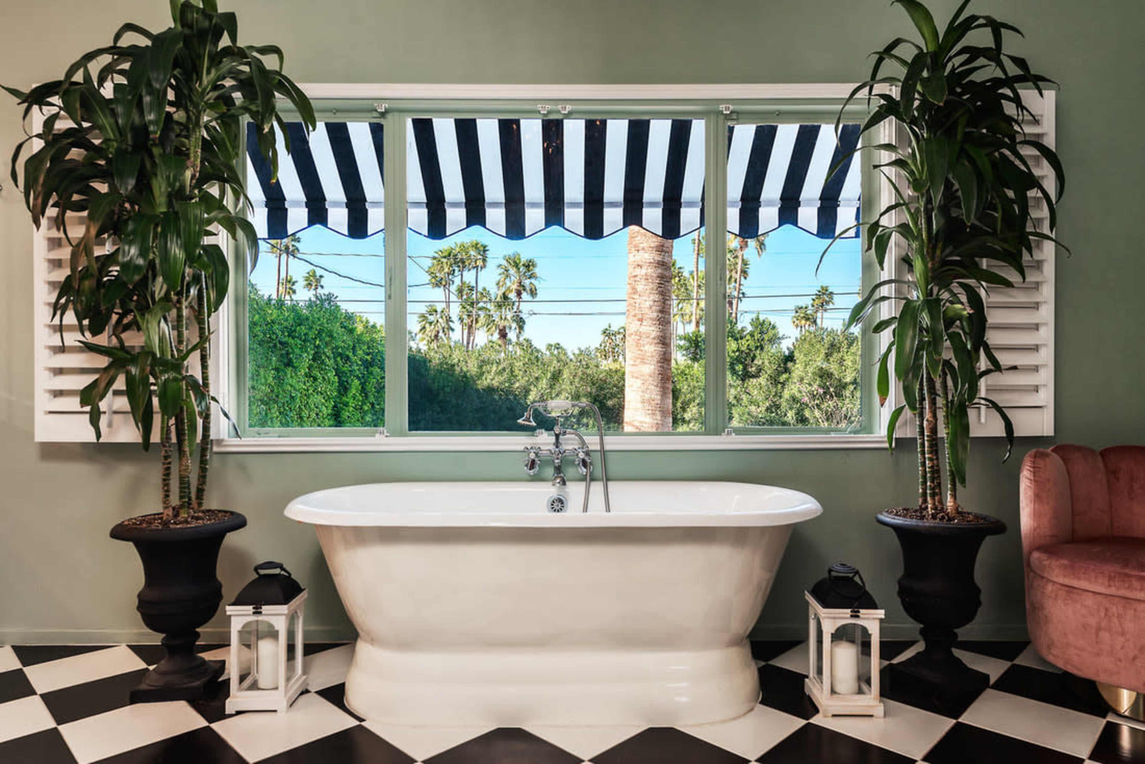 A freestanding white bathtub is positioned between large windows adorned with black and white striped awnings, framed by potted plants, with a checkerboard floor below.
