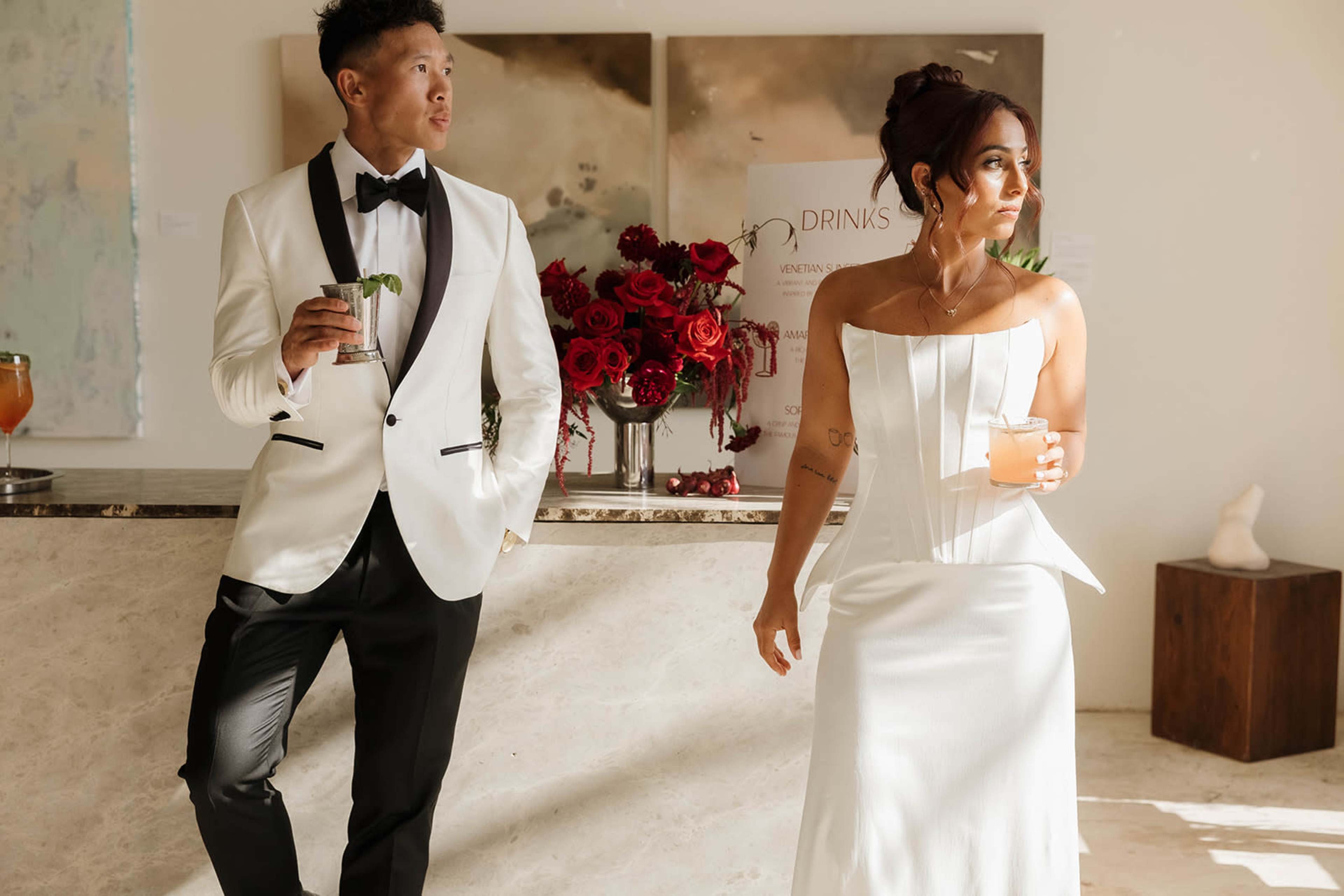 A man in a white tuxedo and a woman in a white dress stand by a bar holding cocktails in a modern, elegantly decorated space.