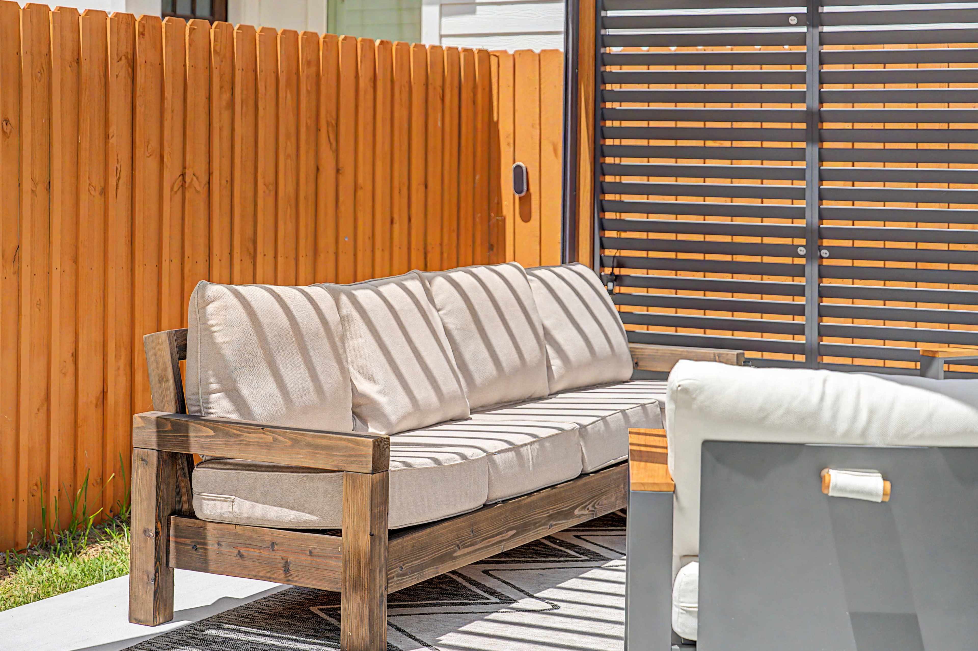 A wooden outdoor sofa with beige cushions is positioned in a patio area near a wooden fence and a shaded structure.