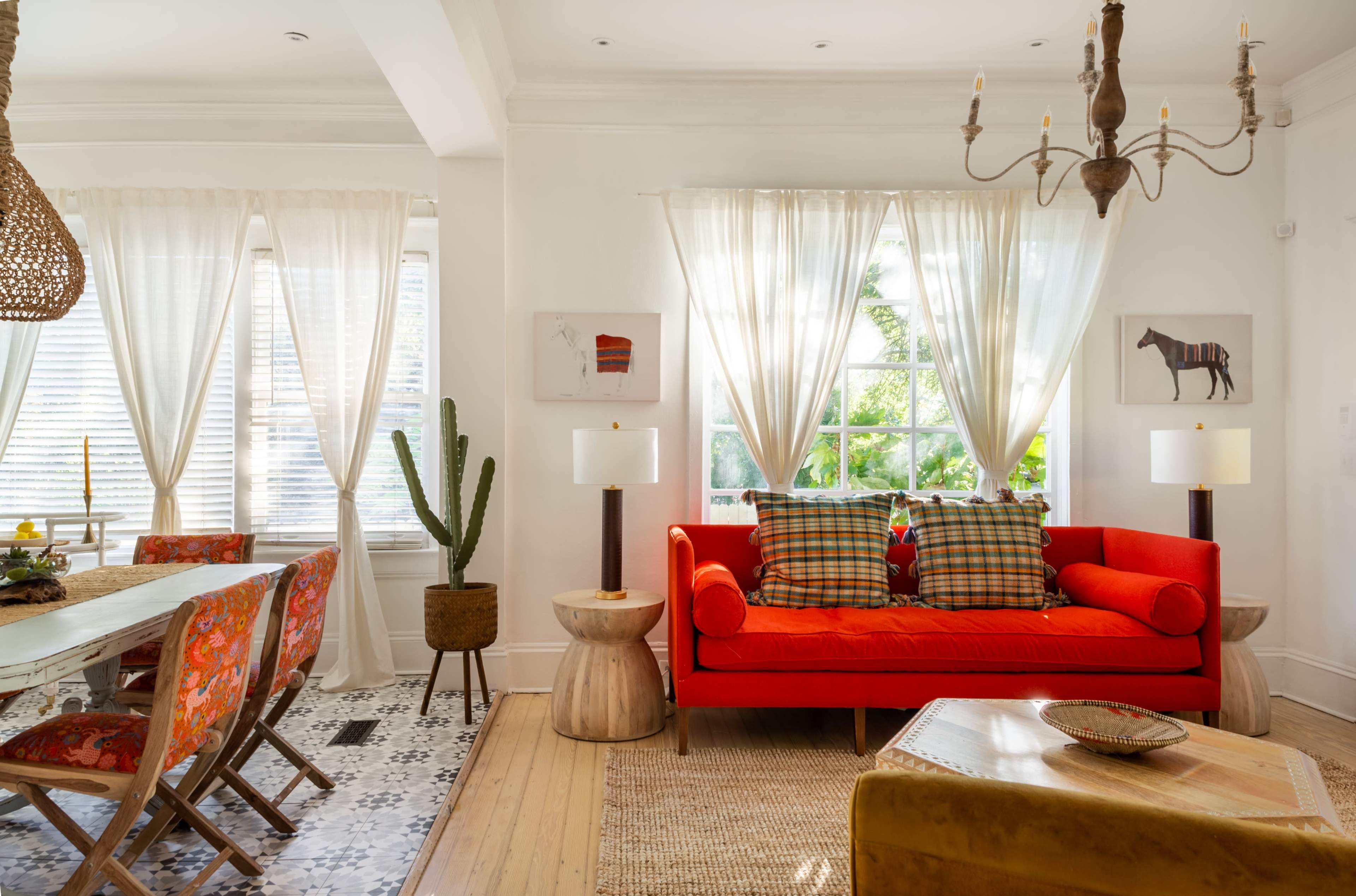 A bright living space featuring a red sofa, two side tables, a dining area with colorful chairs, and large windows adorned with sheer curtains.