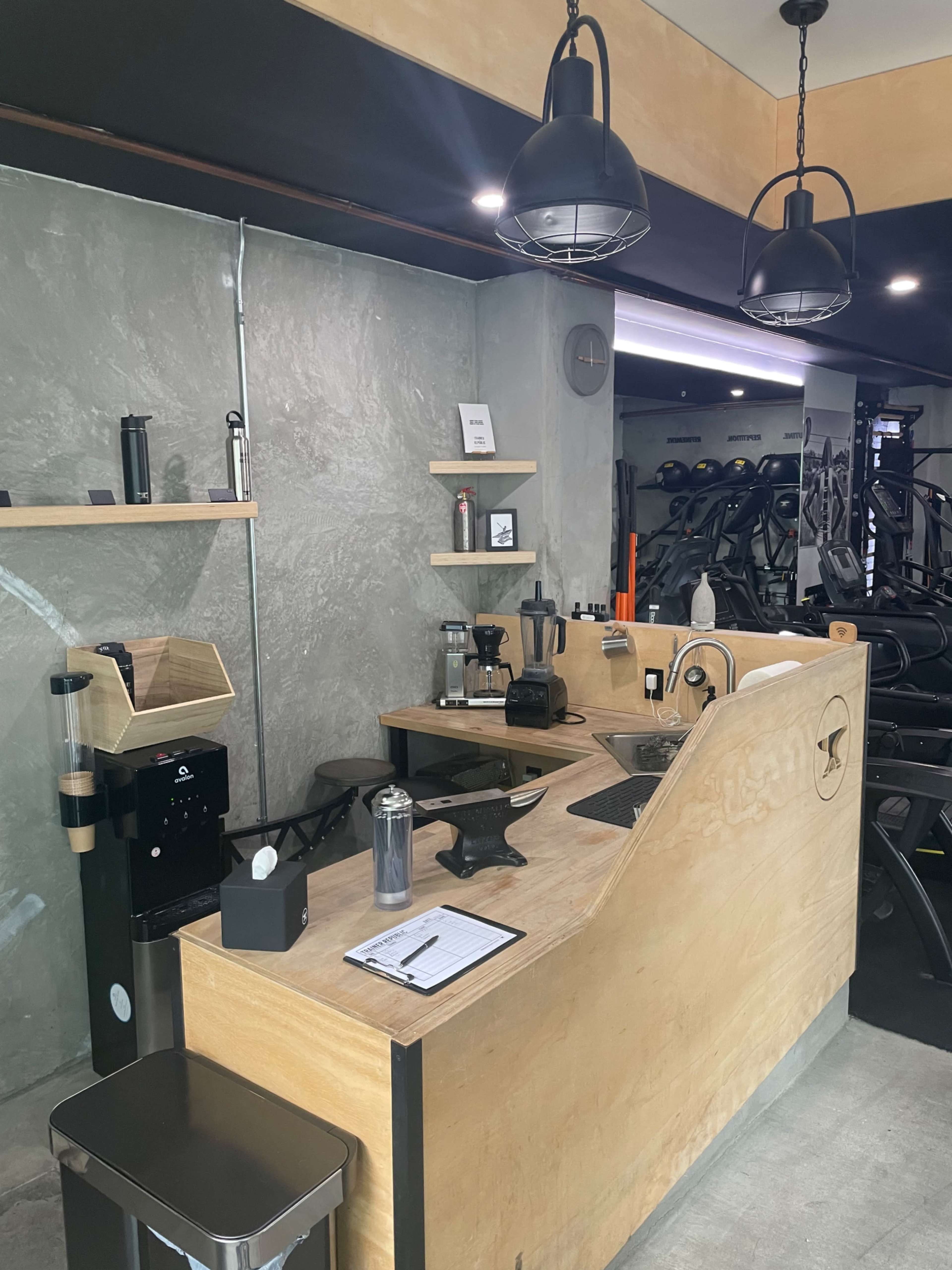 A modern coffee station set against a textured wall in a gym environment, featuring various coffee-making equipment and a wooden counter.