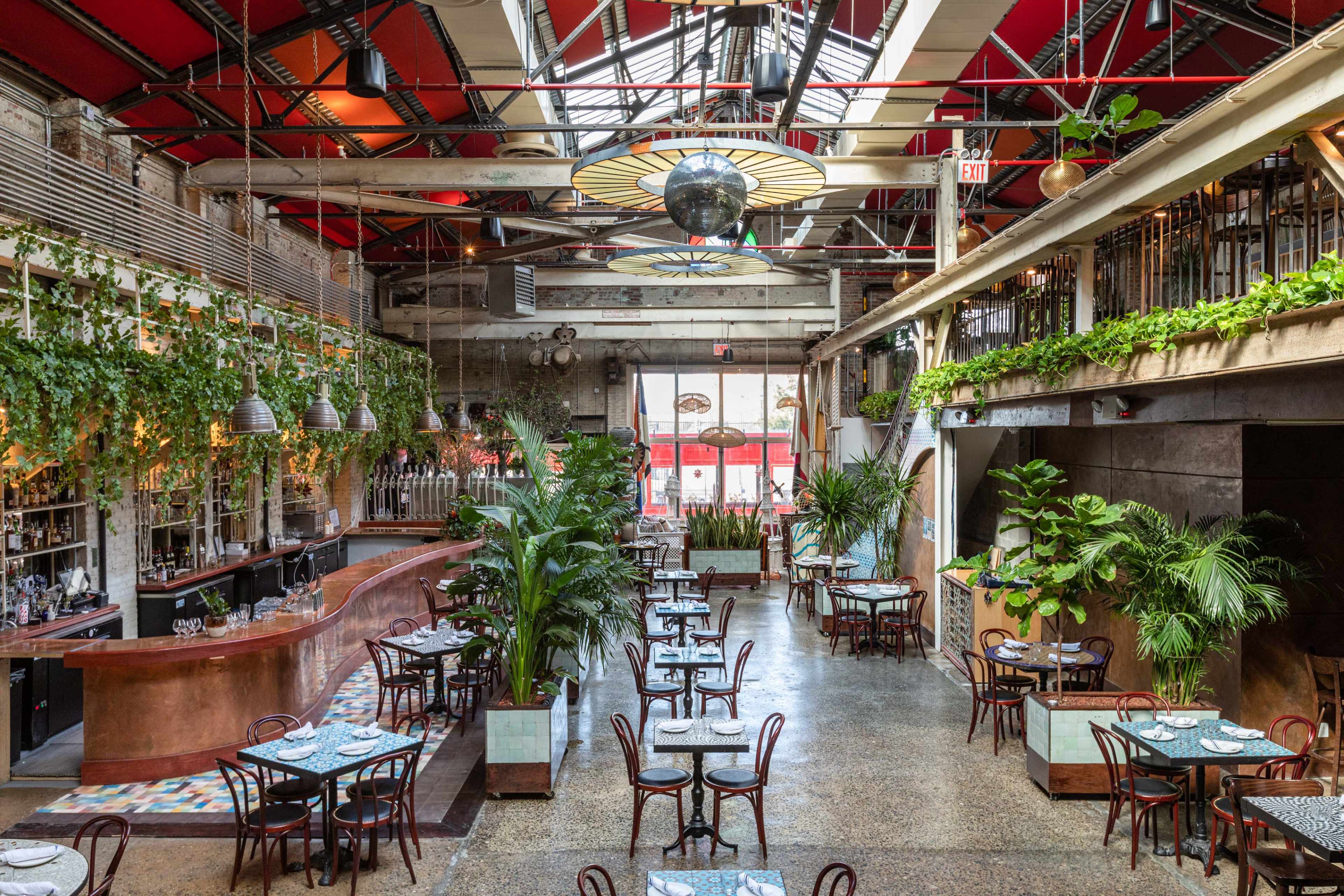 A spacious restaurant with a bar and tables arranged throughout, featuring large plants and overhead lighting.