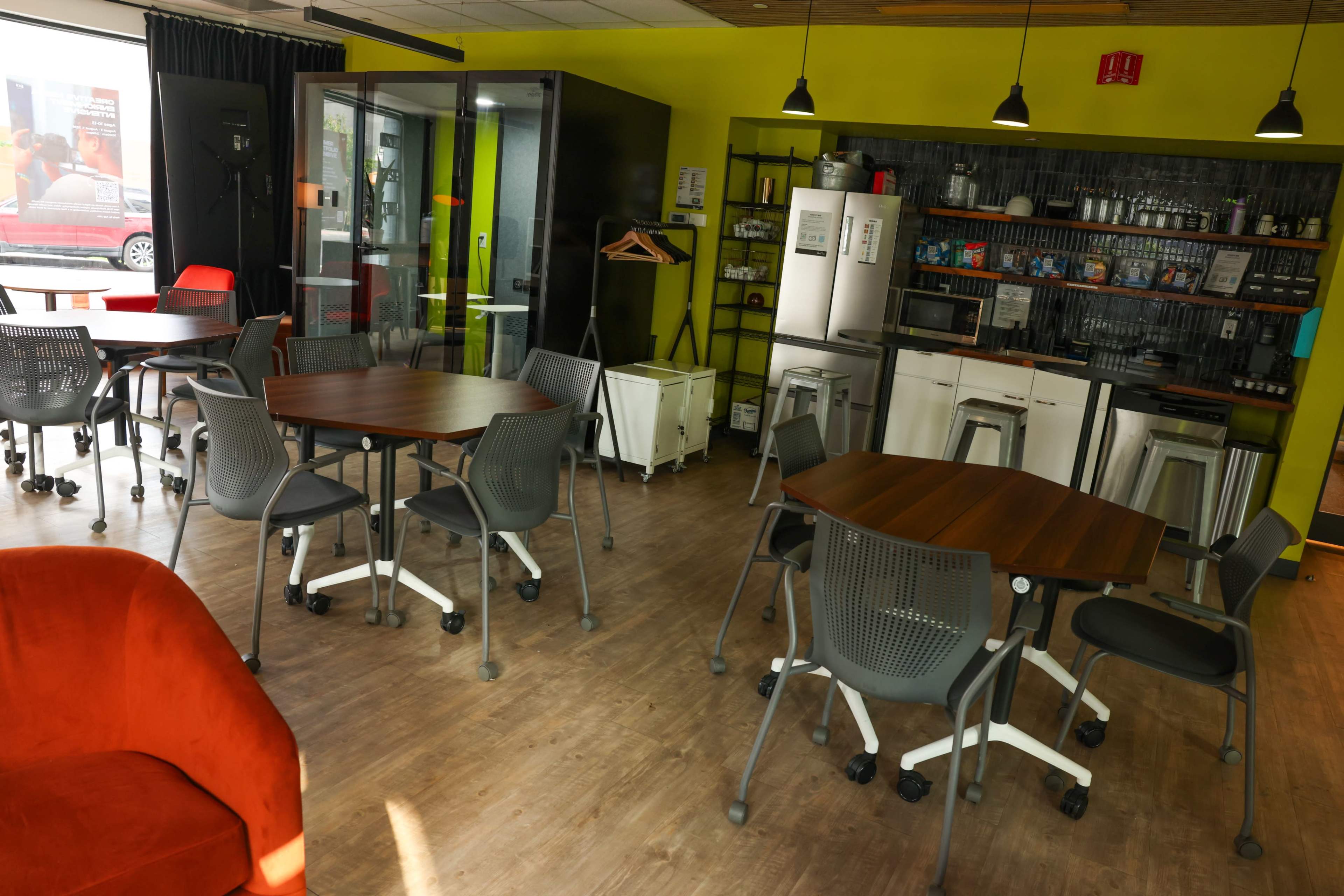 The image shows a modern common area with multiple tables and chairs, a kitchenette, and a glass-walled meeting room.