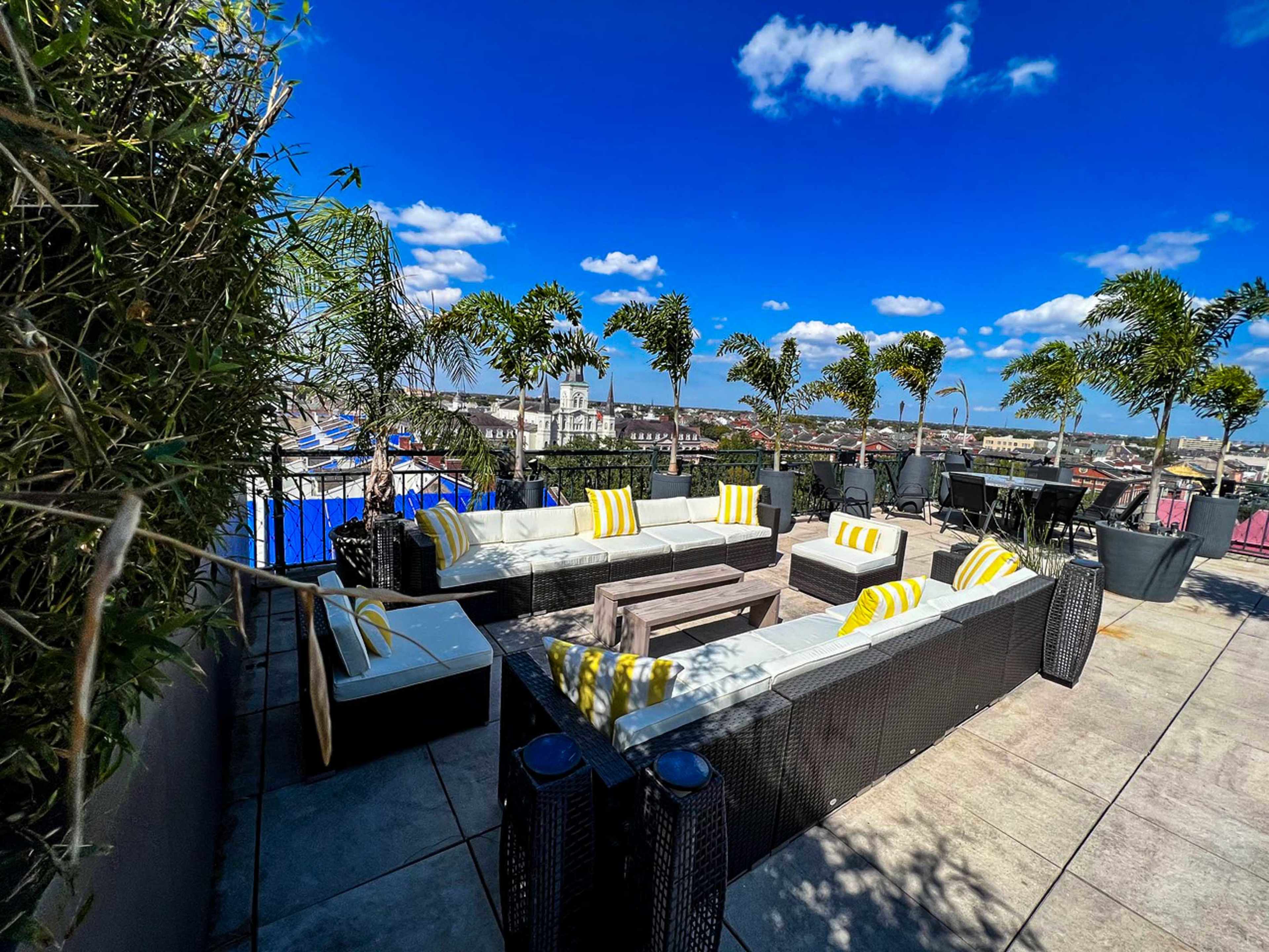 The image shows a rooftop lounge area with modern seating, bright yellow cushions, and palm trees, set against a clear blue sky.
