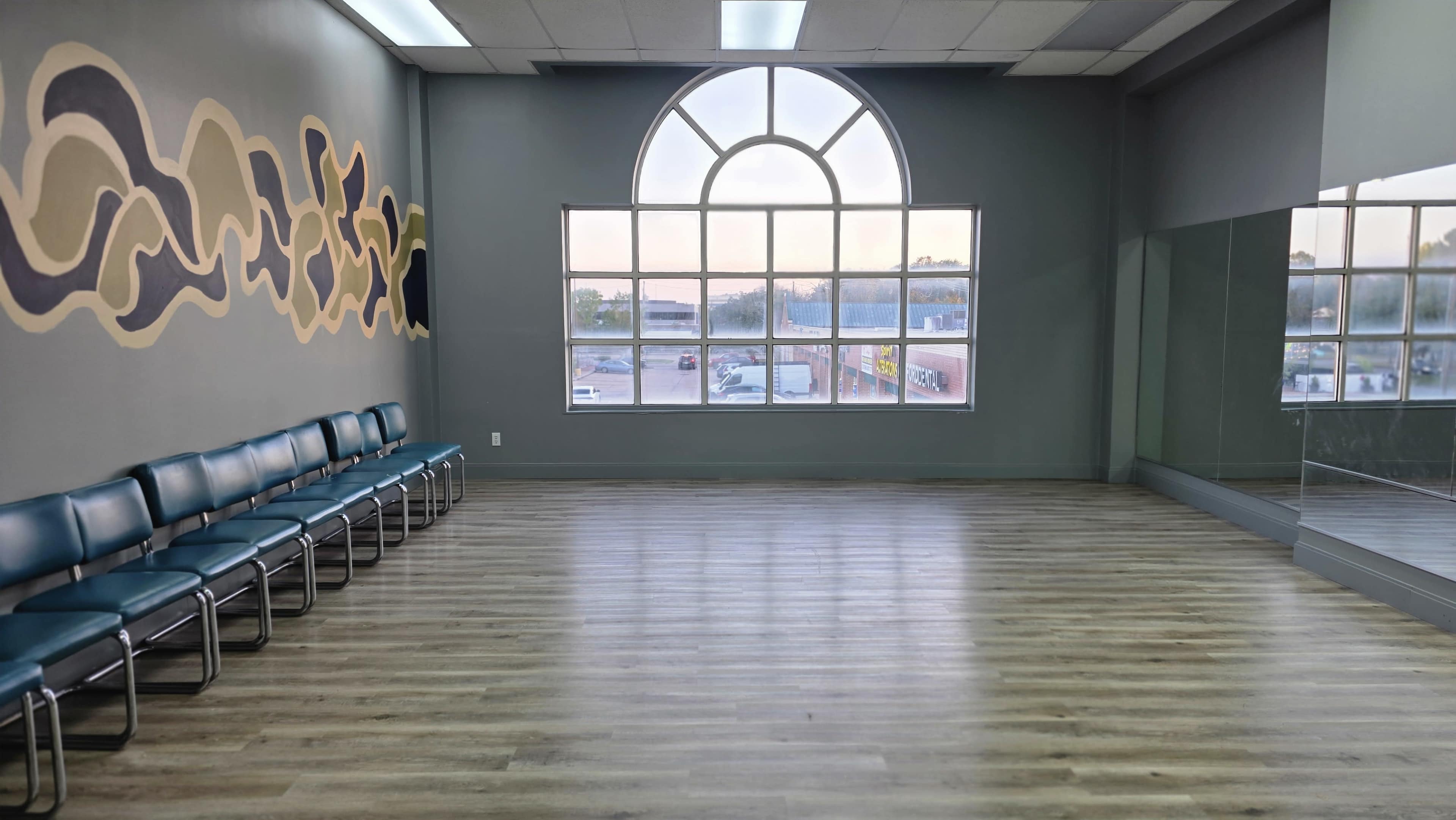 The image shows a spacious, empty room with wooden floors, a large window featuring an arched design, and teal chairs lined up against one wall, with abstract artwork on another wall.