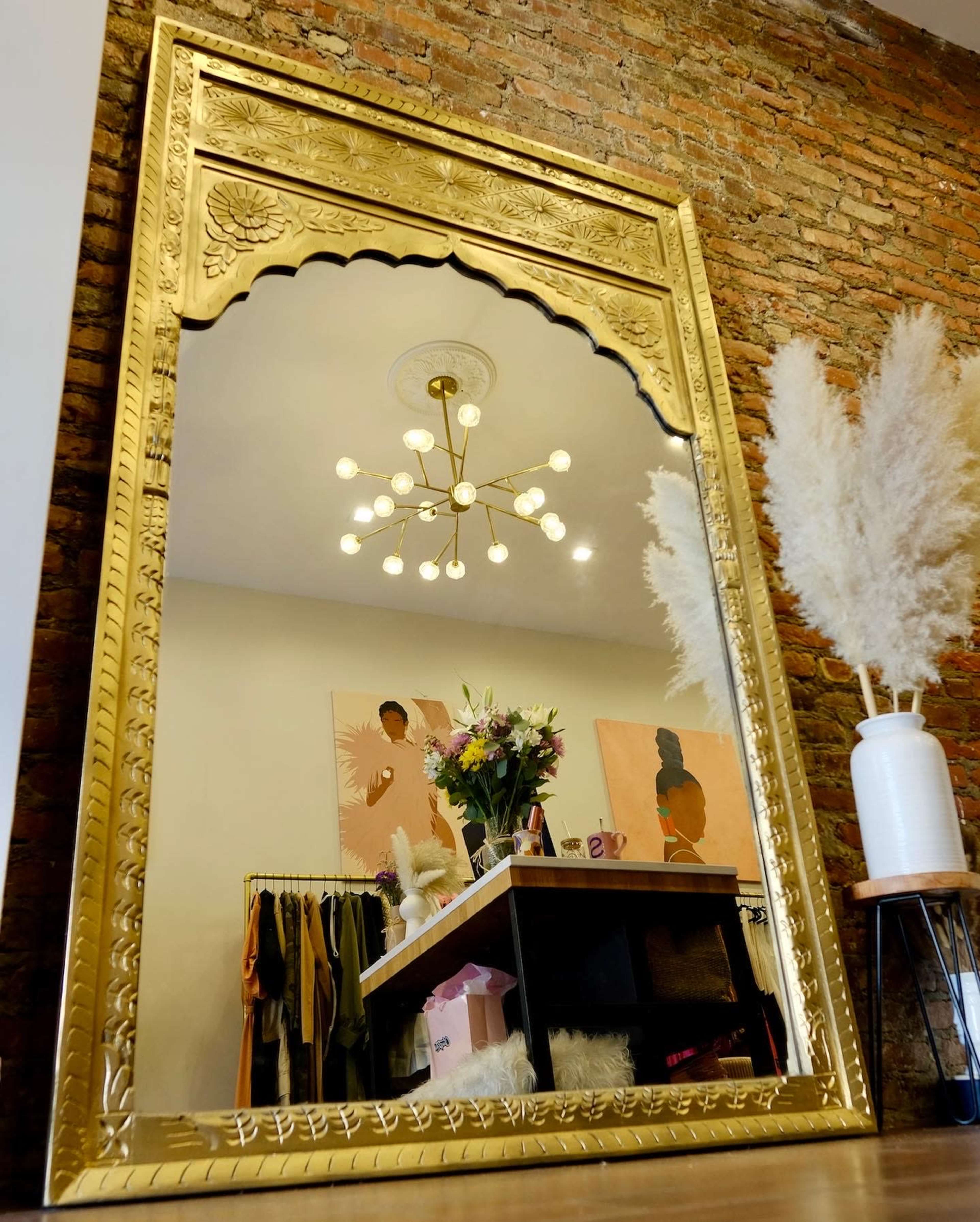 A large ornate gold mirror reflects a room with a clothing rack, floral arrangements, and a modern light fixture.